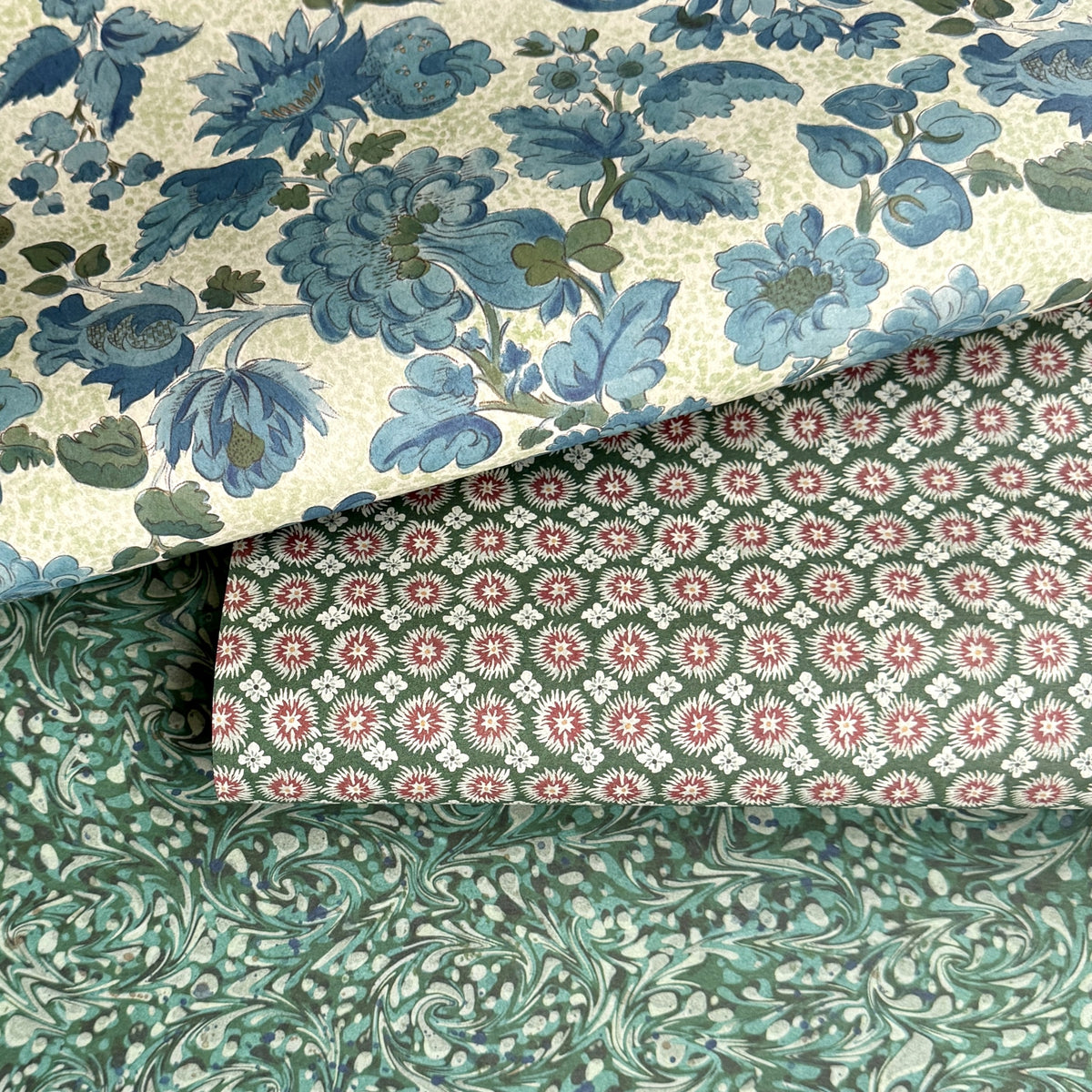 Three overlapping wrapping paper sheets with distinct patterns: a cream background with large blue floral motifs, a dark green background with repeating pink and white starburst medallions, and a teal-green background with an abstract swirling marbled pattern.