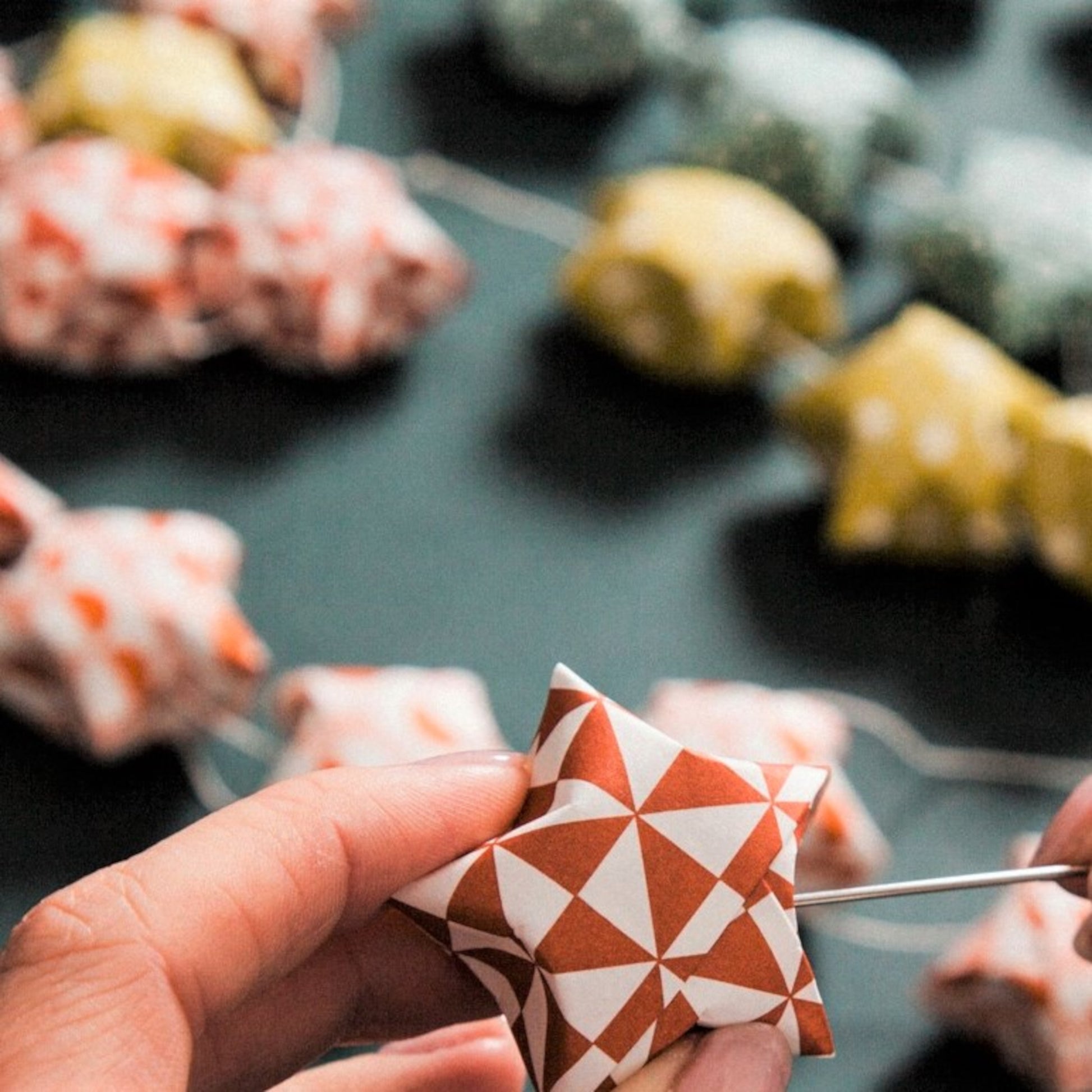 A garland of small paper origami patterned stars "lucky stars" in mustard, teal, aqua and orange, close-up of an orange star being attached to the garland string