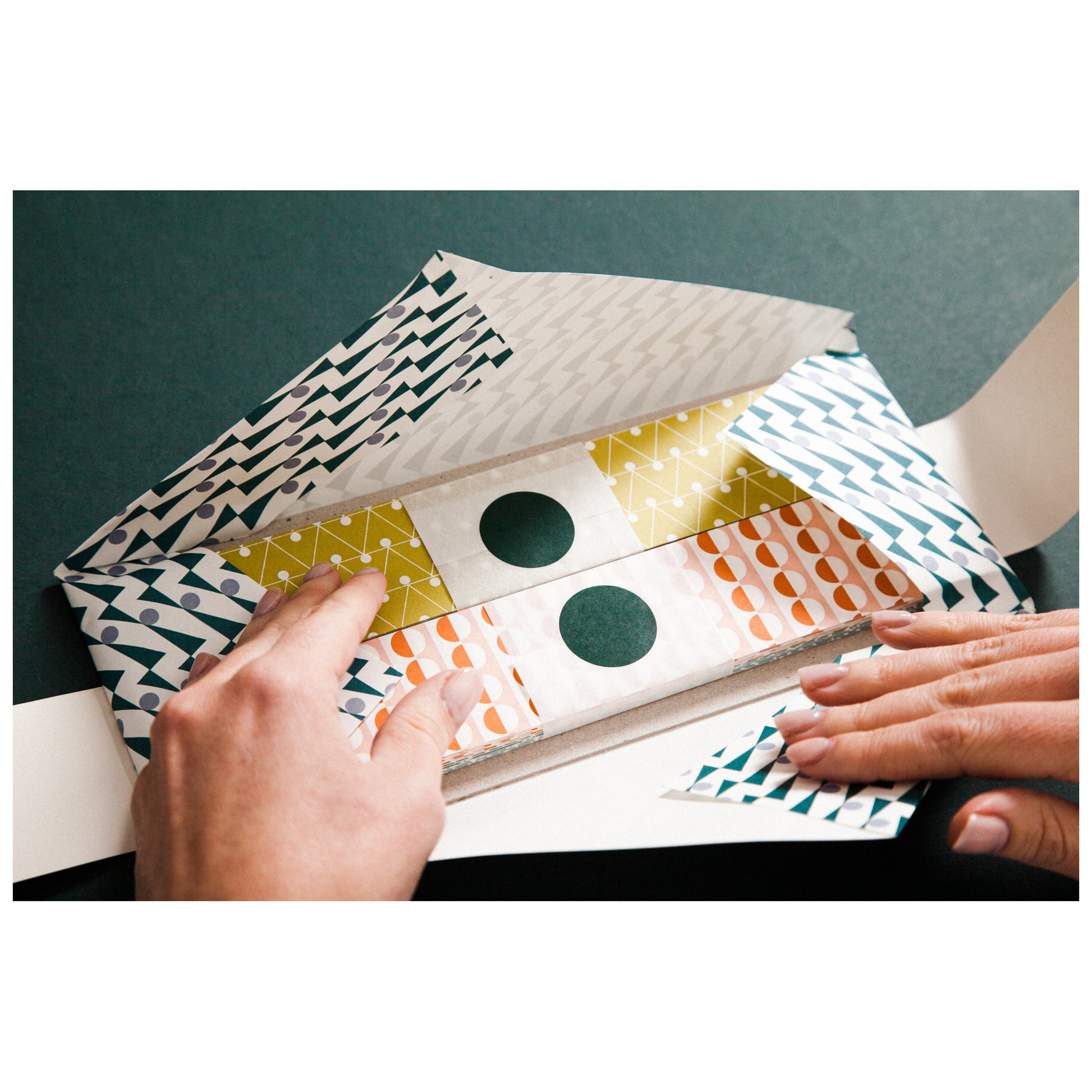 A paperchain of different patterned geometric papers in colours of mustard, orange, aqua and teal, showing the outer envelope being opened to reveal the paper strips inside