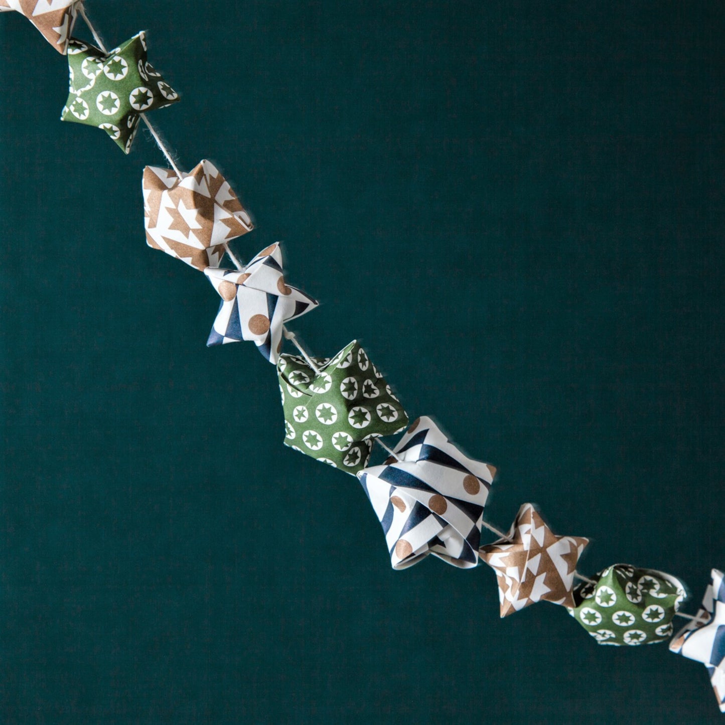 A garland of small paper origami patterned stars "lucky stars" in green, gold and dark blue, pictured against a dark backdrop