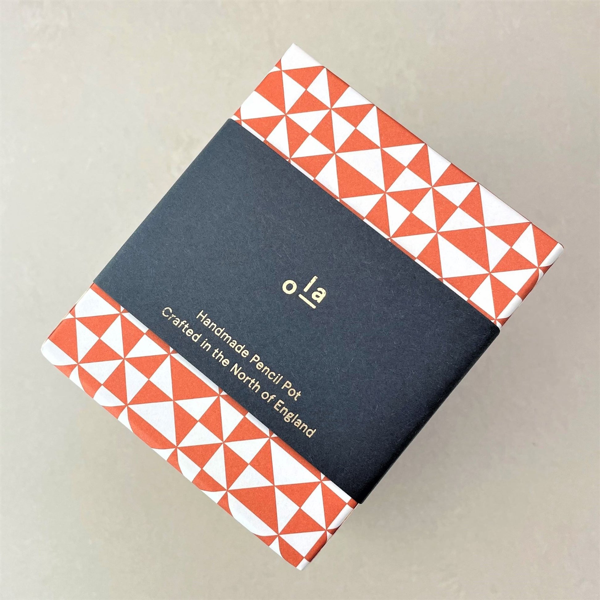 Square pencil pot covered in geometric triangular brick red and white patterned paper, pictured with branded belly band