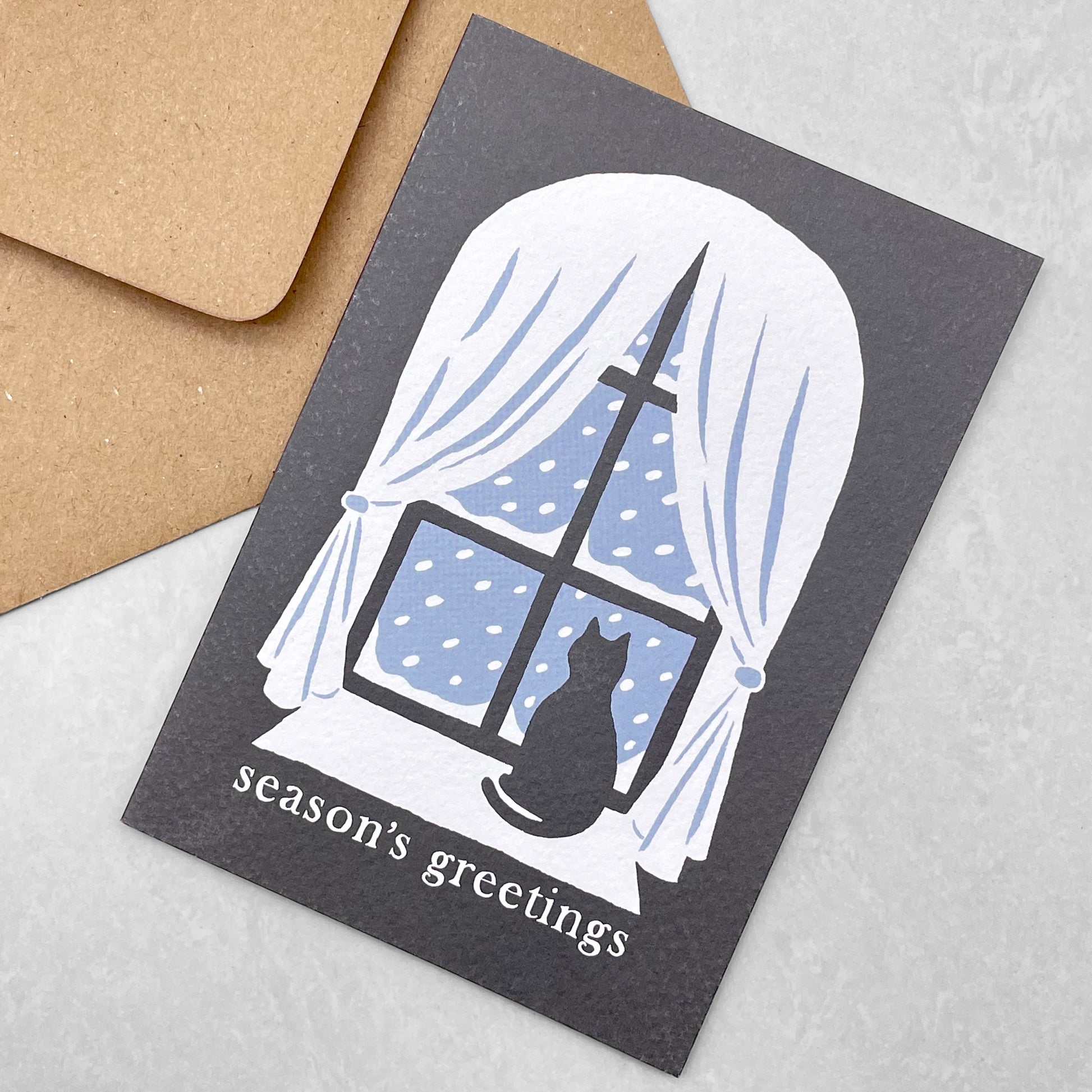 Season's greetings greetings card with a black cat looking out of a window at the snow, by Ariana Martin