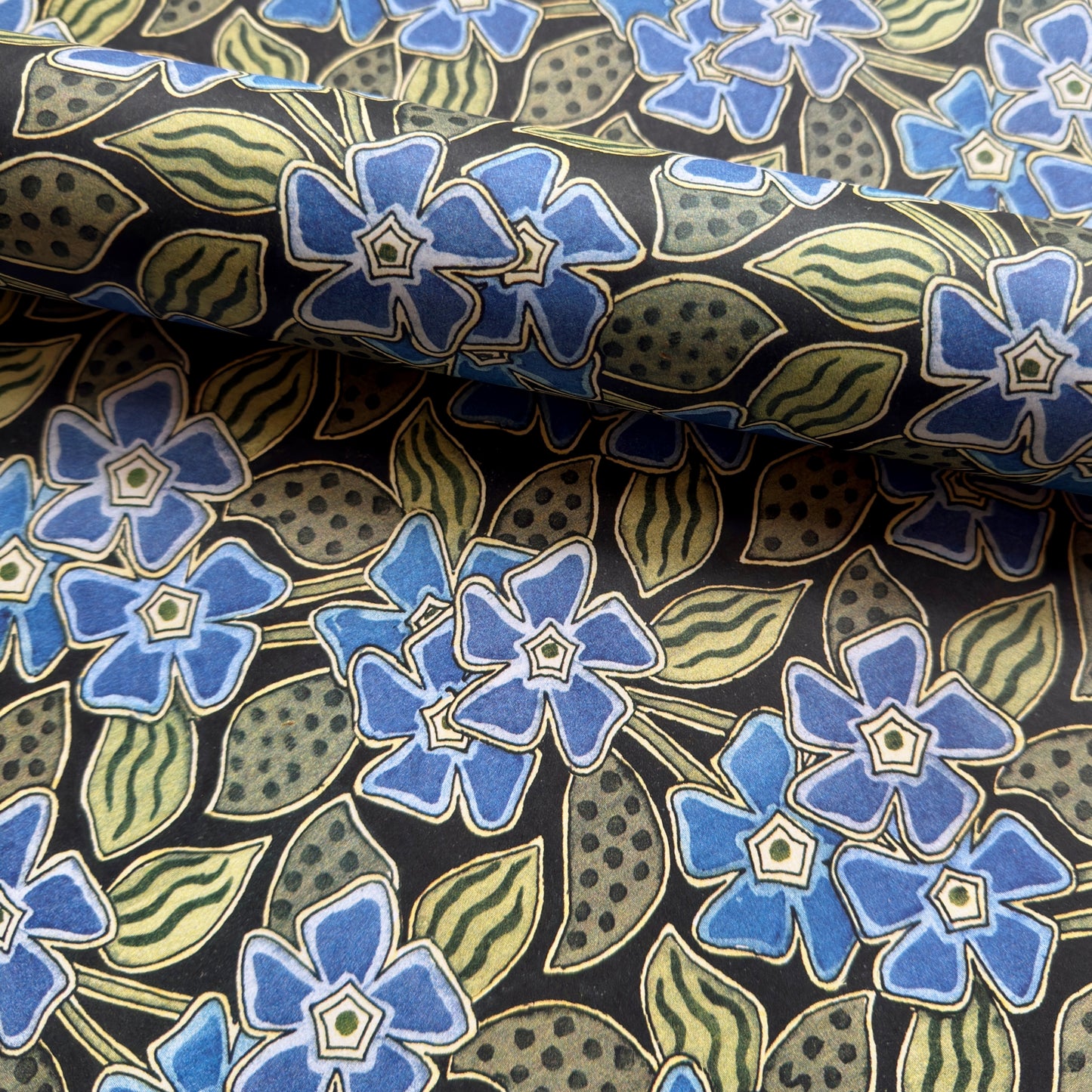 a sheet of wrapping paper with a stylised periwinkle flower design in blue and green on black, close up
