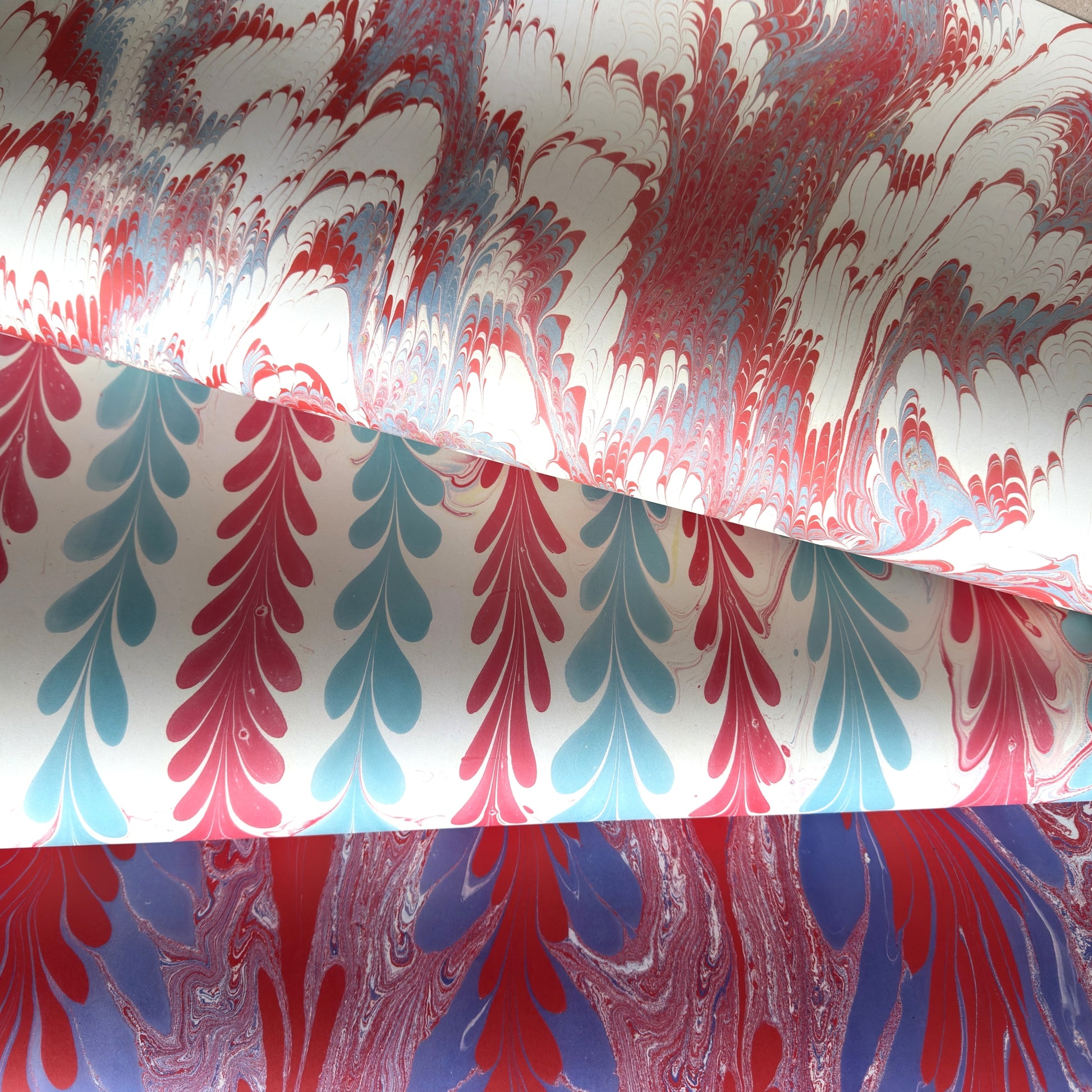 Three rolls of marbled paper with distinct patterns: a red, white, and blue leaf pattern; a red, white, and light blue pattern; and a red, white, and purple marbled pattern, all on a white background.