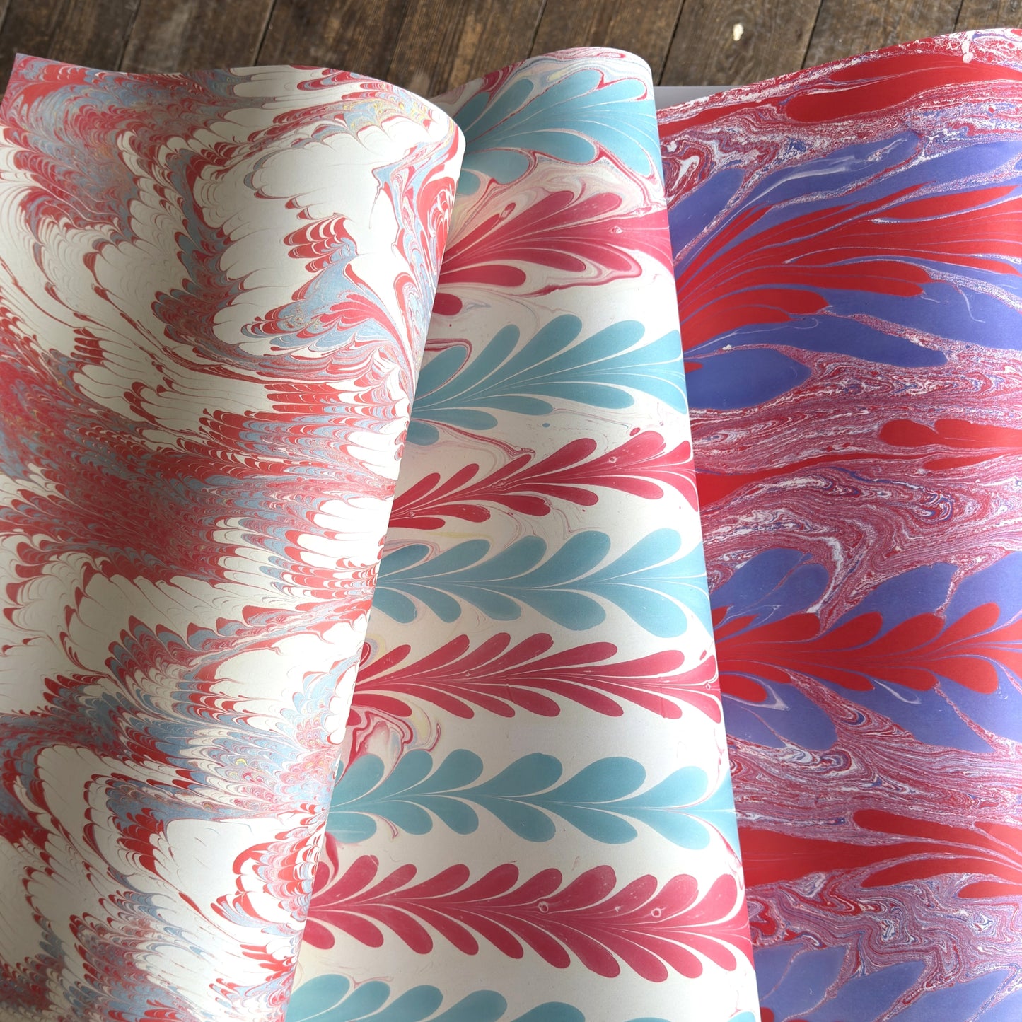 Three rolls of marbled paper with different color combinations of red, blue, and pink swirls on white backgrounds, displayed on a dark wood surface.