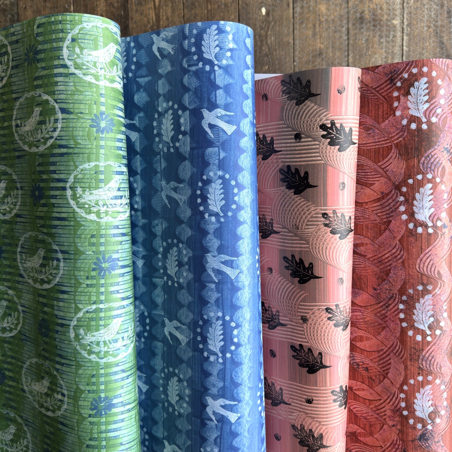 Four rolls of patterned wrapping paper on a wooden surface, showing green, blue, pink, and rust-orange designs with leaf and wave motifs.