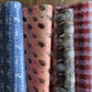 Four rolls of patterned wrapping paper standing upright side by side, showing different color and design options: blue with white bird and leaf motifs, dusty pink with black leaf prints, black with gold wave and globe patterns, and dark red with pink and blue scallop motifs.