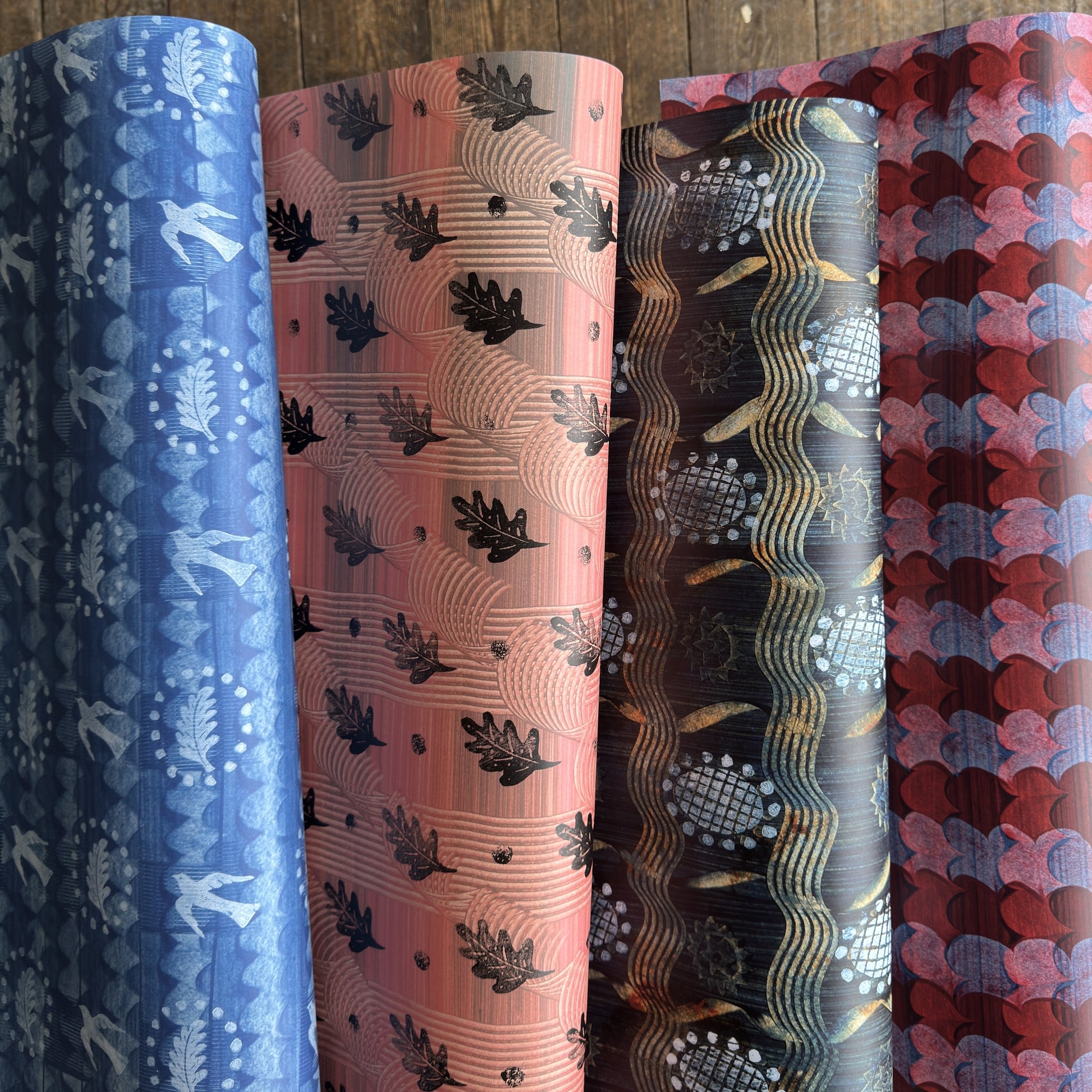Four rolls of patterned wrapping paper standing upright side by side, showing different color and design options: blue with white bird and leaf motifs, dusty pink with black leaf prints, black with gold wave and globe patterns, and dark red with pink and blue scallop motifs.
