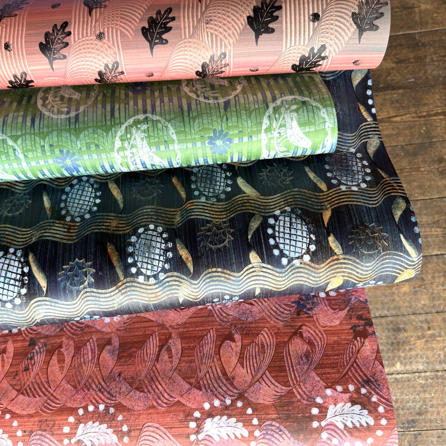 Four sheets of patterned paper stacked diagonally on a wooden floor. The fabrics feature traditional Japanese wave and floral motifs in various colors including pink, green, black, and rust red.