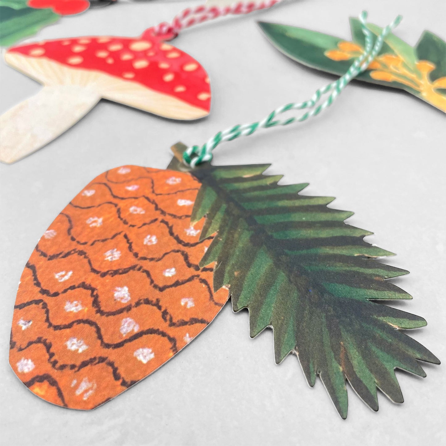 A set of five different festive plant gift tags in red, green and yellow with colourful sttring, by Hadley Paper Goods, pictured a pine cone