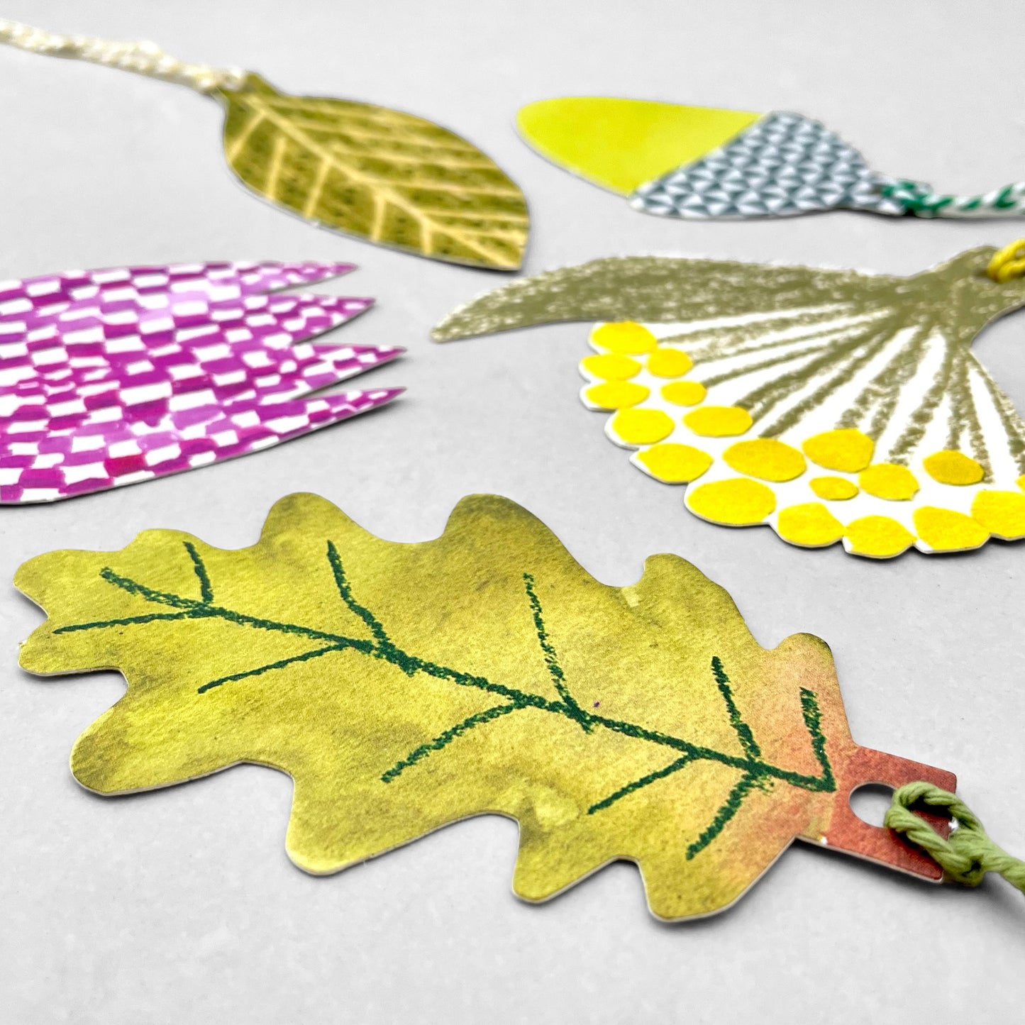 A set of five different plant shaped gift tags in colours of green, yellow and purple, with colourful string by Hadley Paper Goods, pictured an oak leaf