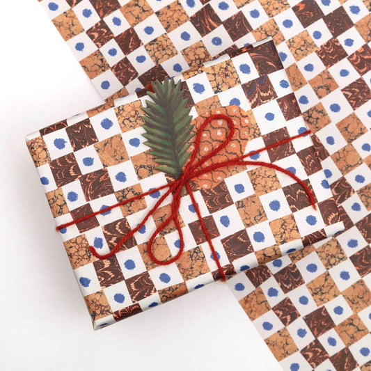 A sheet of wrapping paper by Hadley Paper Goods with a chequerboard pattern of brown marbled squares on a white background with a blue dot.  Pictured wrapped as a present with a tag