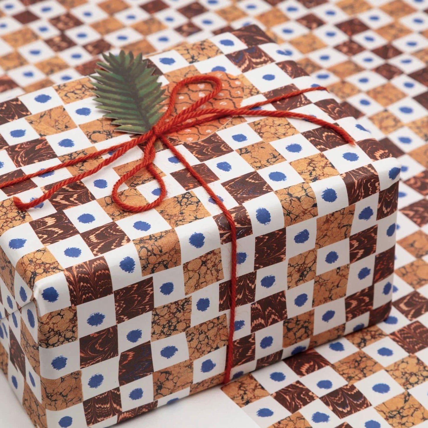 A sheet of wrapping paper by Hadley Paper Goods with a chequerboard pattern of brown marbled squares on a white background with a blue dot. Pictured wrapped as a present with a red wool ribbon and tag