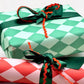 a sheet of wrapping paper with a two tone green large diamond pattern. By Heather Evelyn and shown wrapped as a gift with green satin ribbon and red rik rac bow