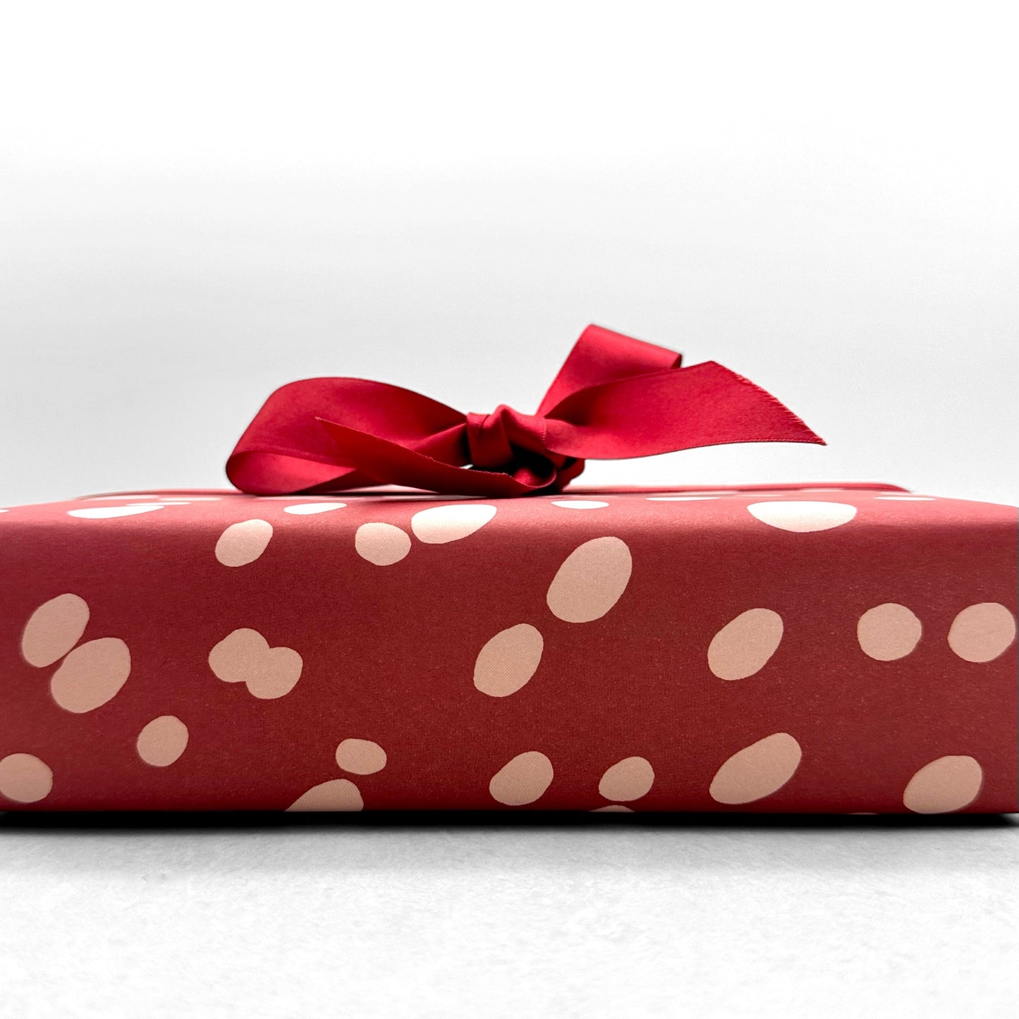 burgundy rouge wrapping paper with random blush pink spots, by Heather Evelyn. Pictured wrapped with a red satin ribbon bow