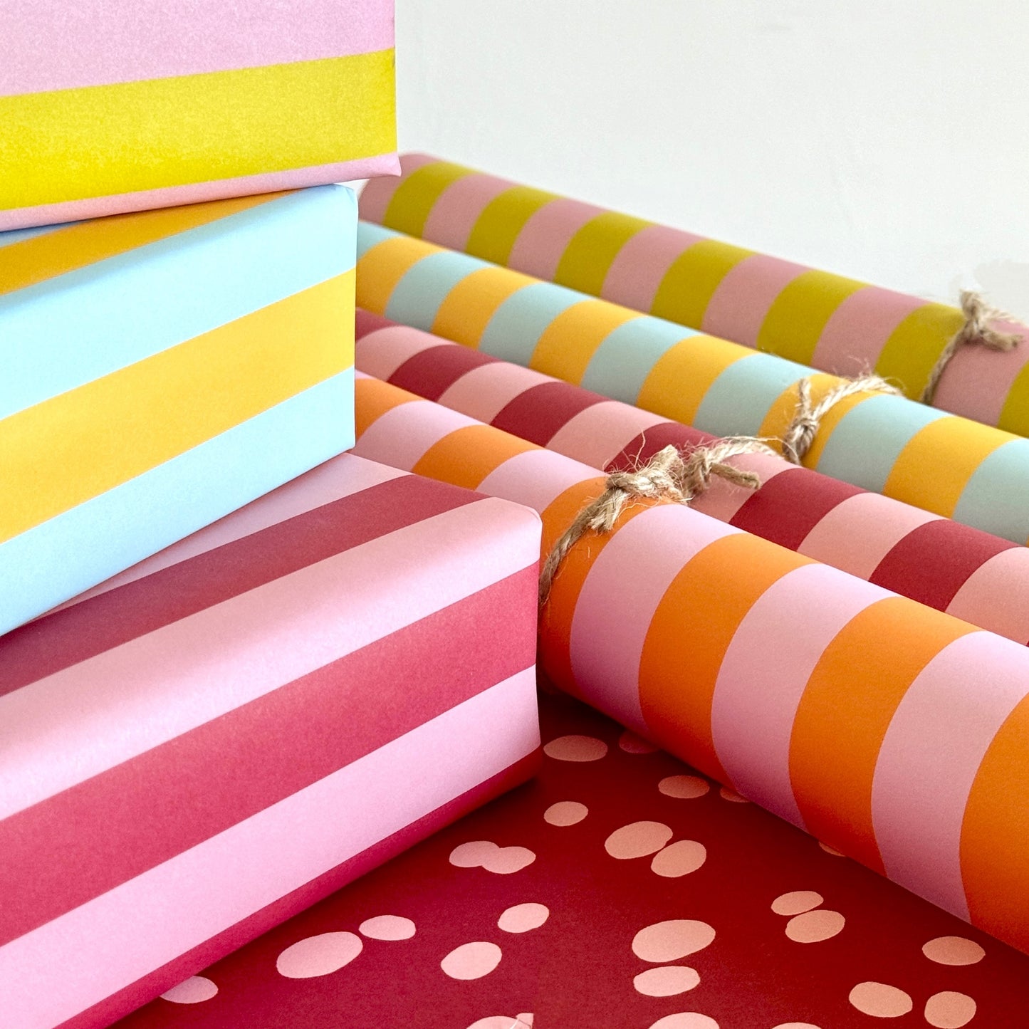 wide classic stripe wrapping paper in orange and pink by Heather Evelyn. Pictured rolled with other striped designs and a pile of presents
