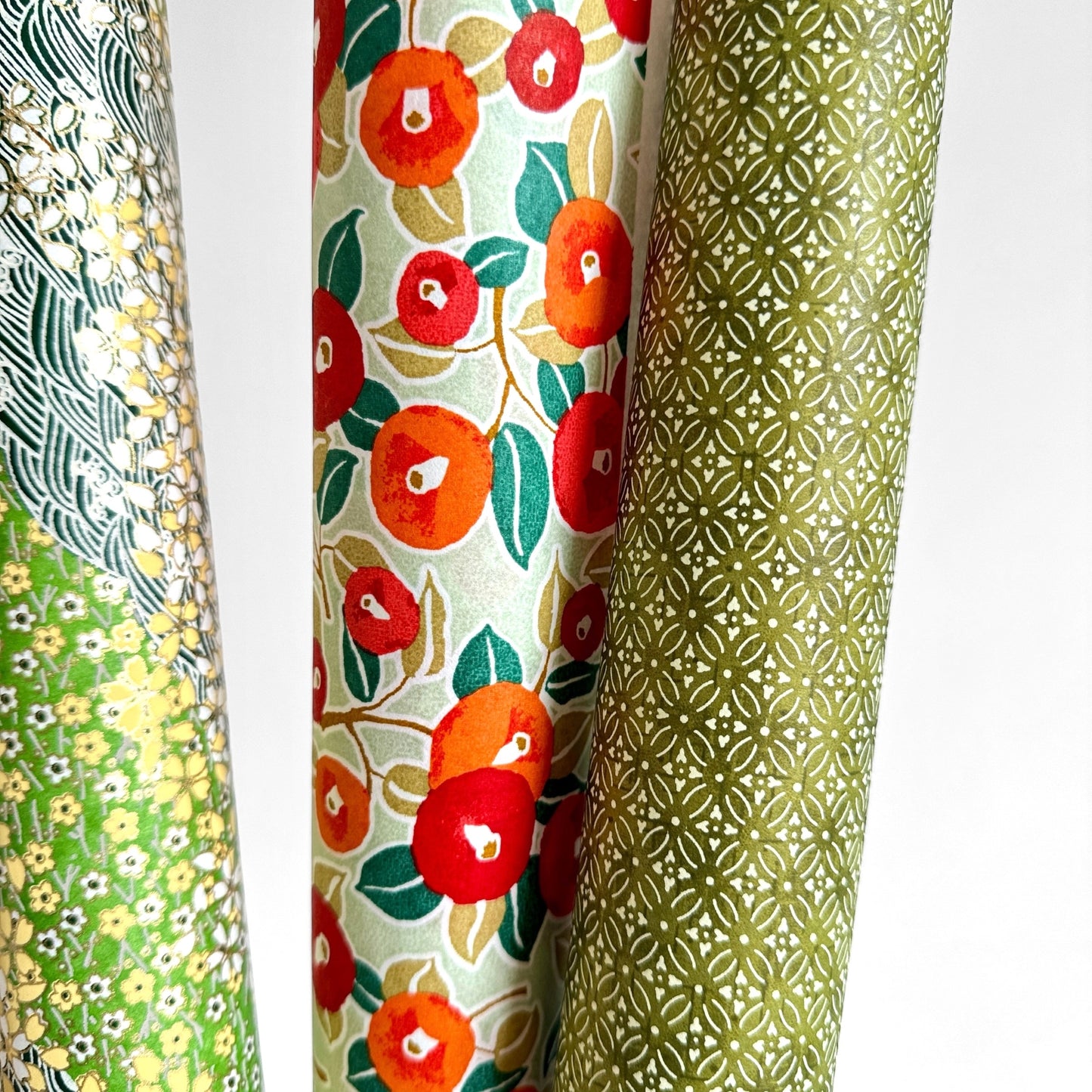 Japanese silkscreen chiyogami paper with a repeat pattern of camellia flowers in red and orange on a soft green ground. Pictured rolled with other designs