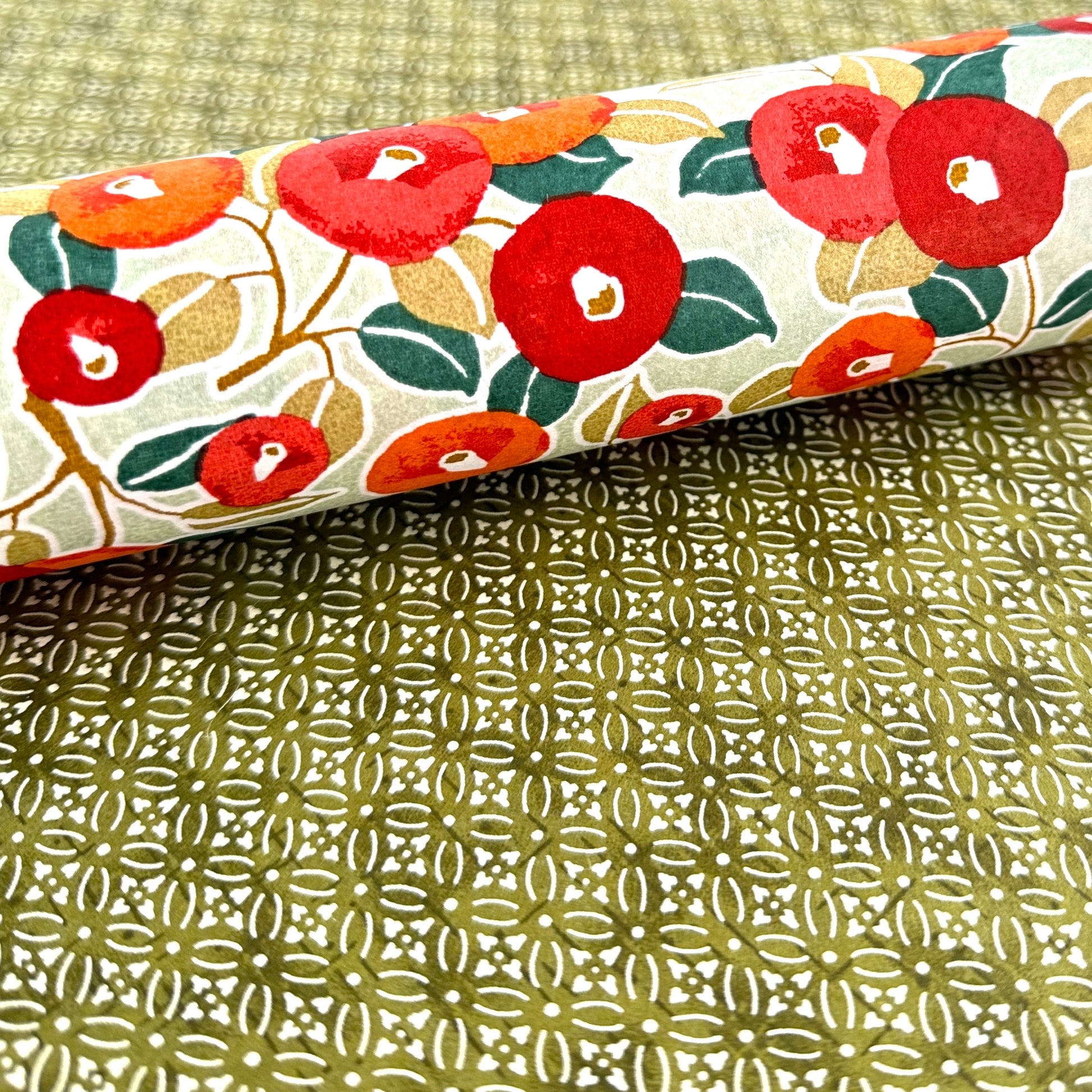 Japanese silkscreen chiyogami paper with a repeat pattern of camellia flowers in red and orange on a soft green ground, close up