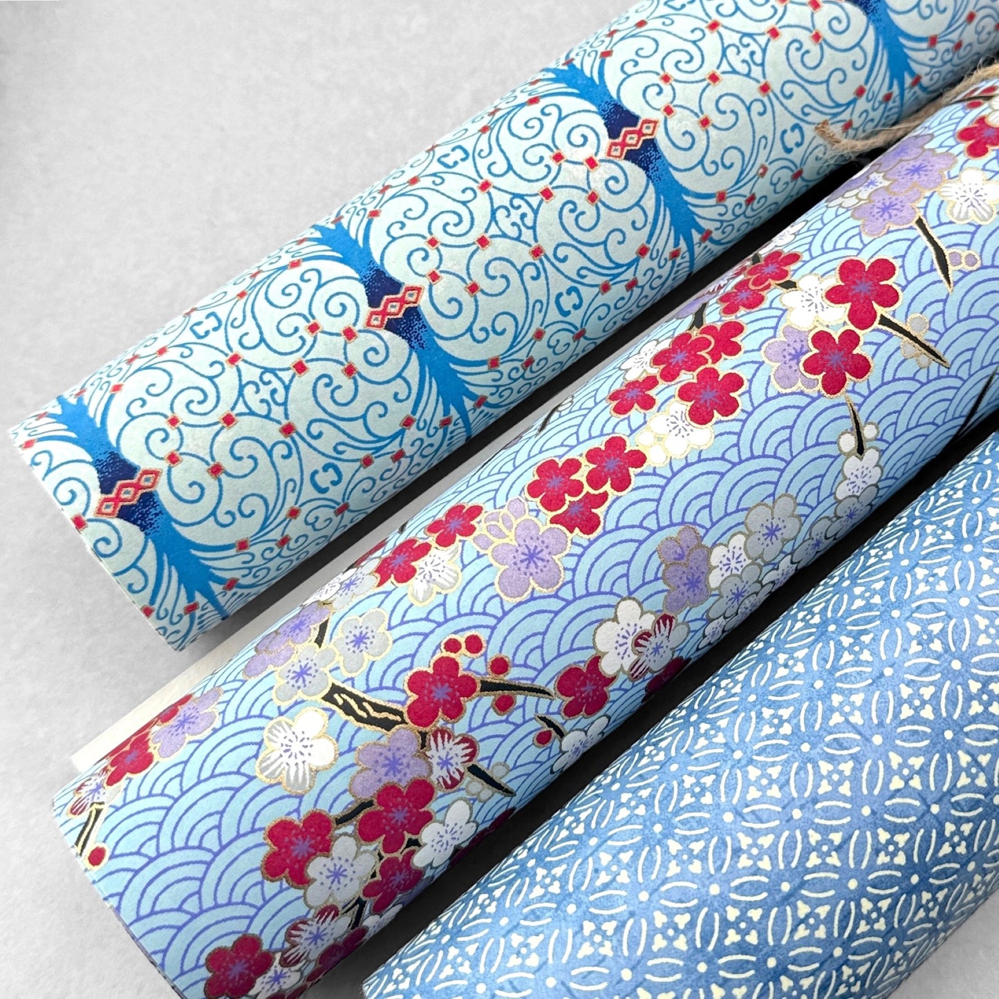 Japanese silkscreen chiyogami paper with a pattern of deep red, lilac and white plum blossom on a blue backdrop, rolled and alongside other designs