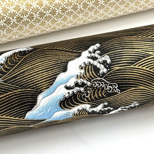 a sheet of Japanese silkscreen printed chiyogami paper with a depiction of pale blue stormy waves in a sea in black and gold. close up