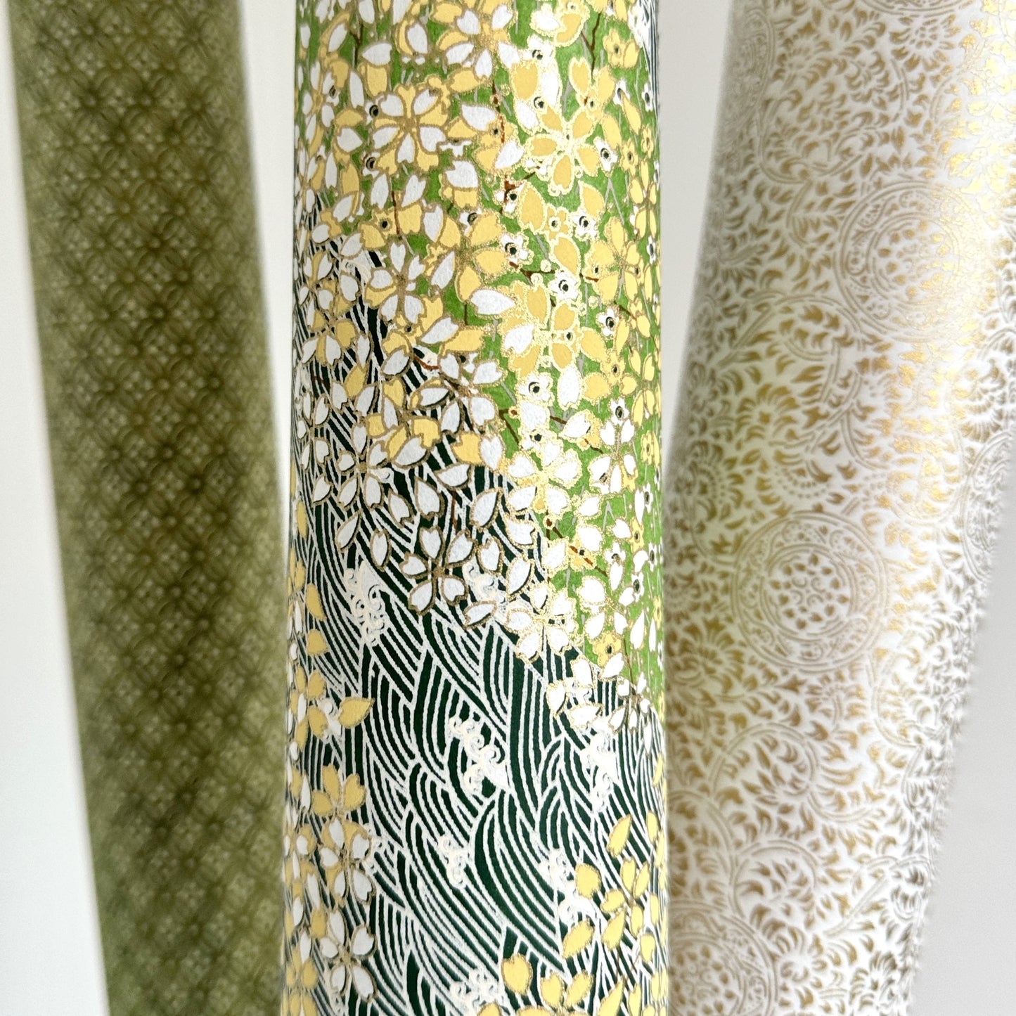 Japanese silkscreen chiyogami paper with a pattern of dark green rivers and grass green banks with an abundance of little flowers in white and yellow. Pictured rolled with other papers