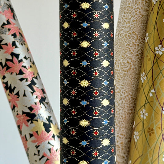 Japanese silkscreen chiyogami paper with a pattern of small repeat motifs in red, blue, lemon and gold on a black background, pictured rolled alongside other designs