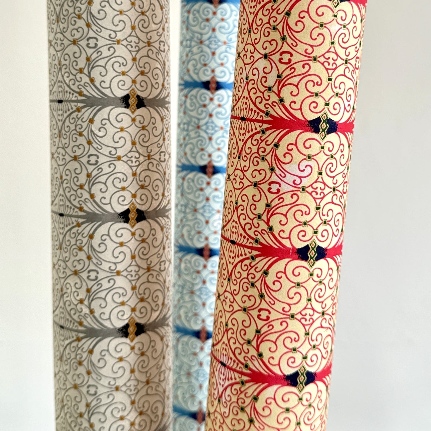 Japanese silkscreen chiyogami paper with a intricate filigree trellis design in pink with gold accent, pictured with rolled sheets of other designs