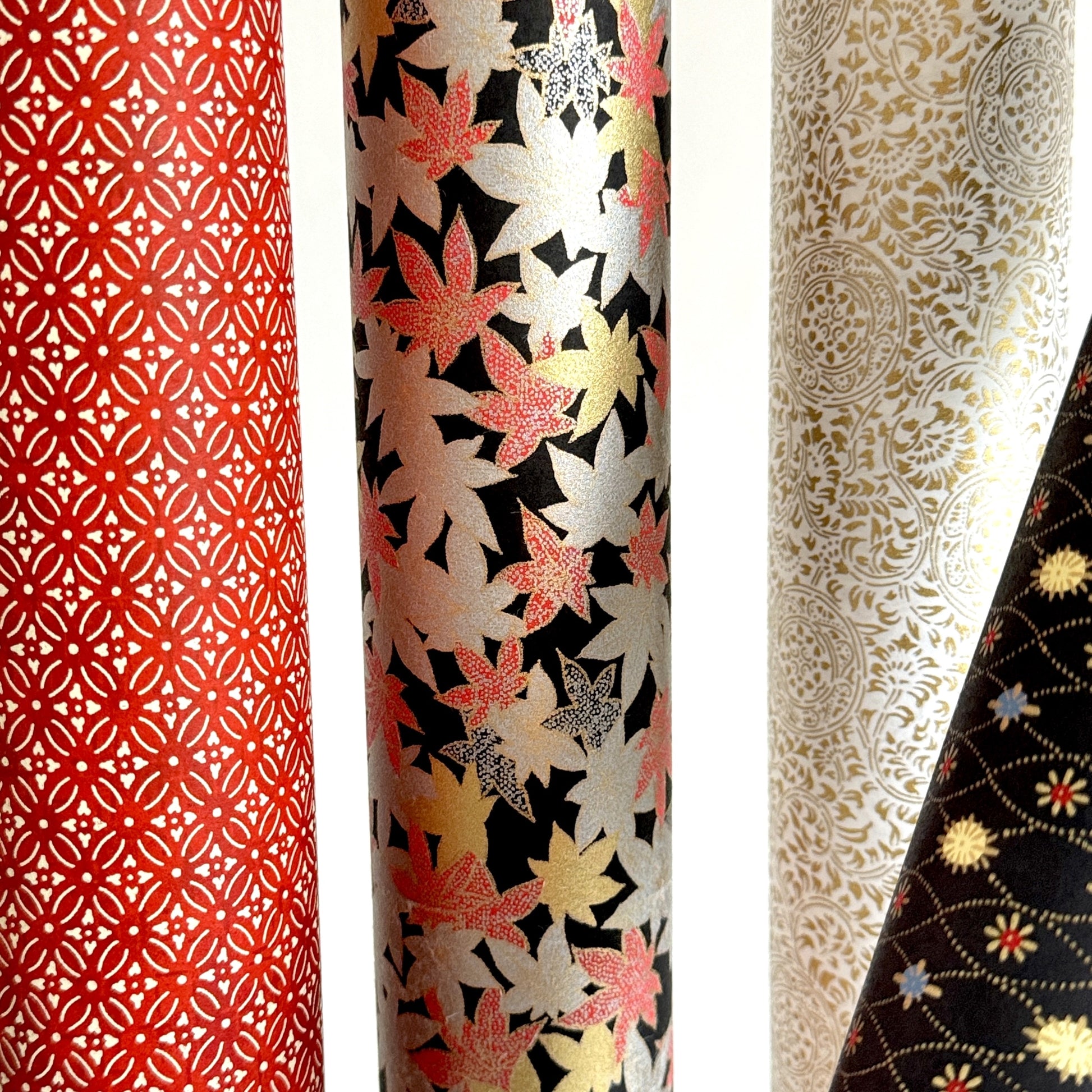 Japanese silkscreen chiyogami paper with an abundance of falling maple leaves in red, gold and silver on black backdrop, rolled alongside other patterns.
