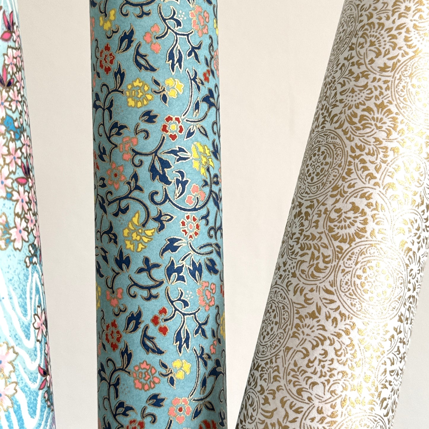 Japanese silkscreen chiyogami paper with a dainty floral pattern in navy, yellow and coral on a teal backdrop. Finished with gold metallic ink. Pictured rolled with other designs.