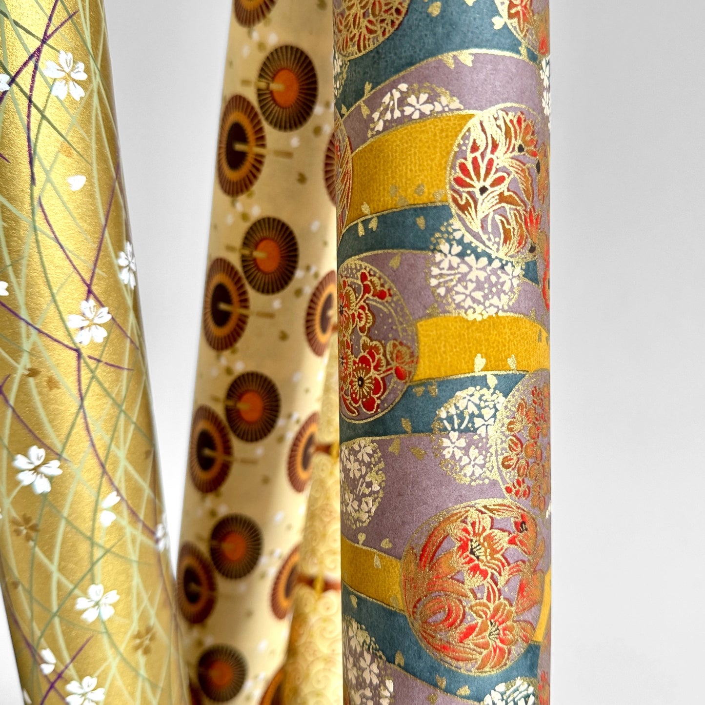 Japanese silkscreen chiyogami paper with a traditional botanical circle motif in tones of mustard, inky blue, lilac and gold. Pictured rolled with other designs