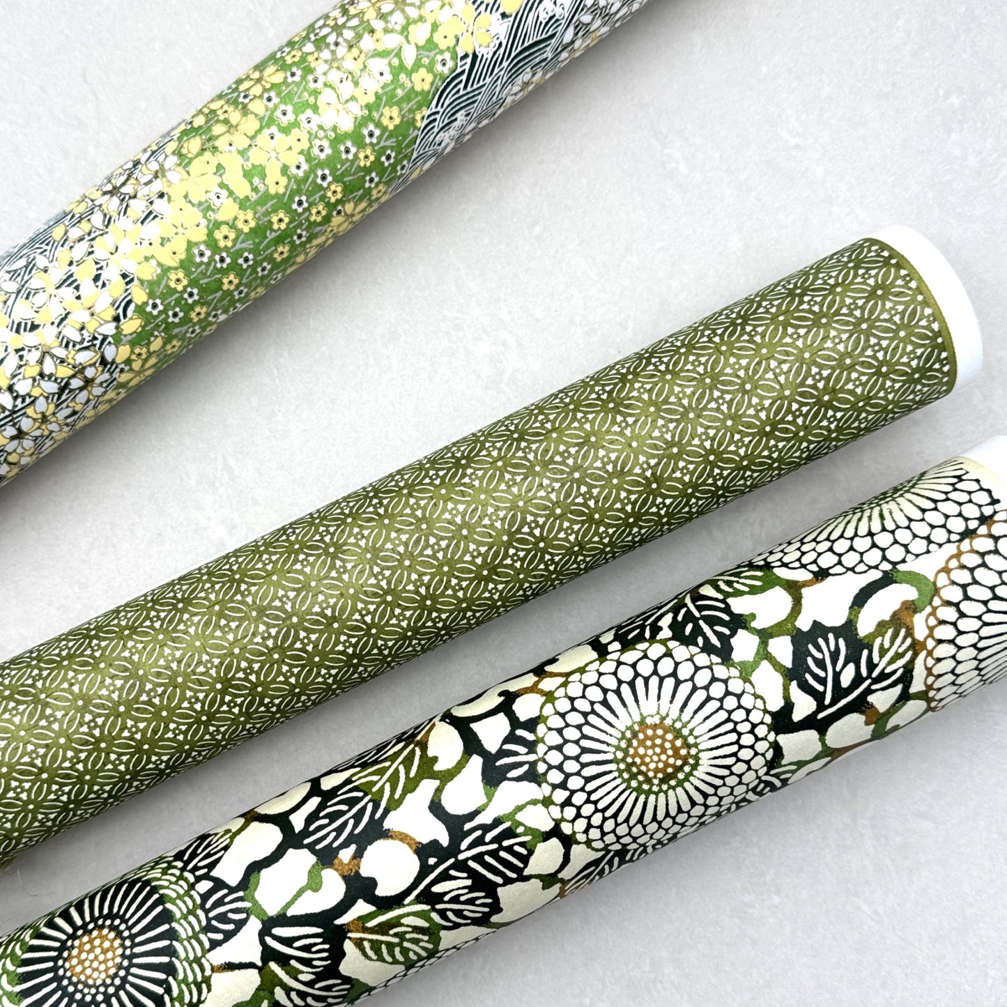 Japanese silkscreen chiyogami paper with a repeat pattern of stylised chrysanthemum flowers in tones of deep green on a buttermilk cream backdrop. Pictured rolled with other green patterned papers