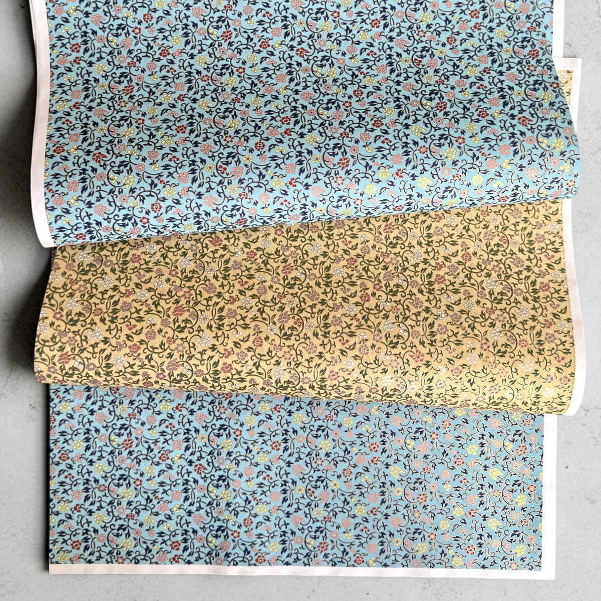 Japanese chiyogami silkscreen paper with a dainty floral pattern on a creamy backdrop and finished with gold metallic ink.  Pictured loosely folded with the same pattern in teal 