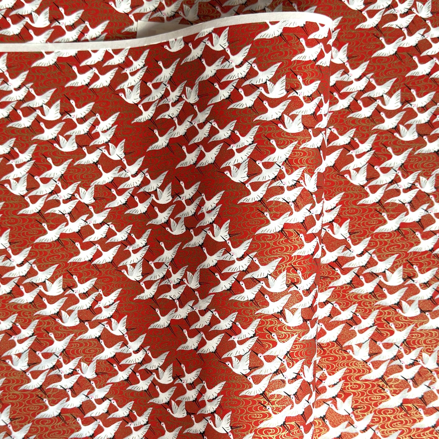 A close-up of a red sheet of Japanese chiyogami paper with an all-over repeating pattern of white cranes and small gold wave-like motifs. 