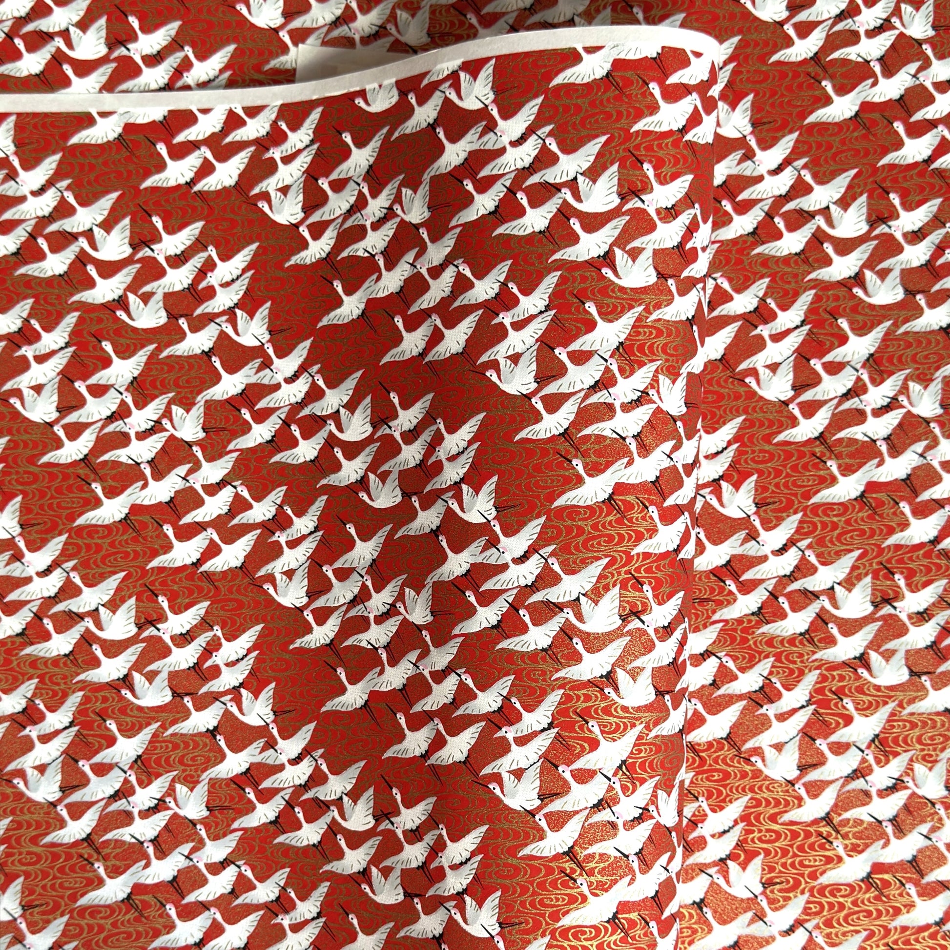A close-up of a red sheet of Japanese chiyogami paper with an all-over repeating pattern of white cranes and small gold wave-like motifs. 