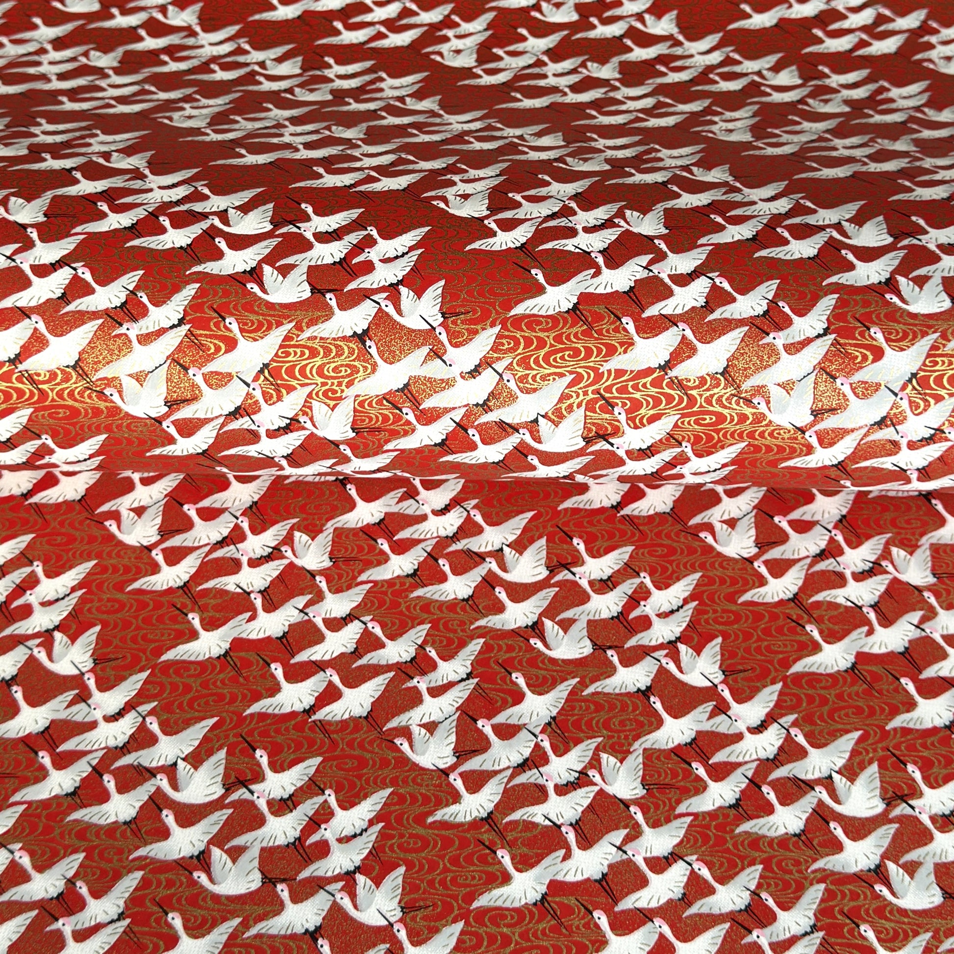 A close-up of a red sheet of Japanese chiyogami paper with an all-over repeating pattern of white cranes and small gold wave-like motifs. 