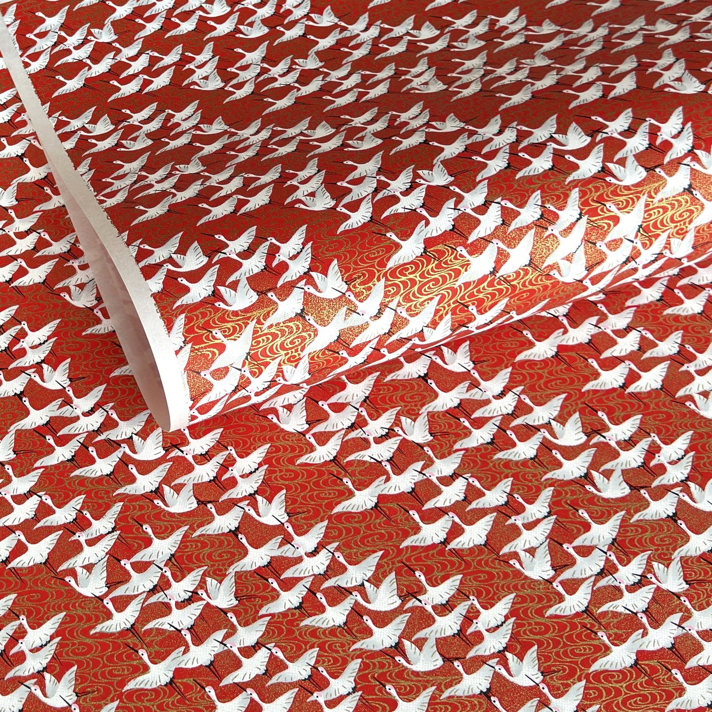 A close-up of a red sheet of Japanese chiyogami paper with an all-over repeating pattern of white cranes and small gold wave-like motifs. 