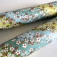 Japanese chiyogami paper silkscreened with a dainty pattern of white sakura flowers on an backdrop of aqua and lime diagonal wavy stripes. Pictured rolled