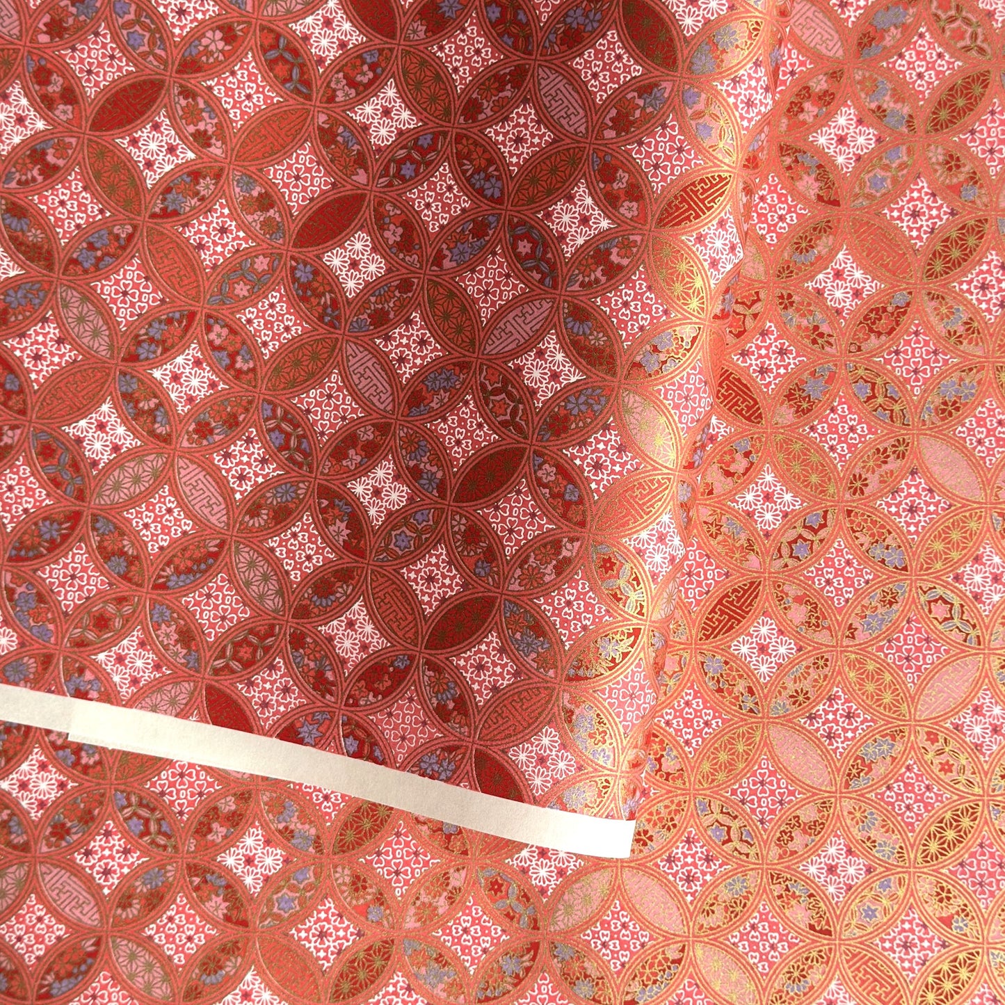 A close-up of a sheet of Japanese chiyogami paper featuring a repeating geometric medallion design in shades of terracotta red, coral pink, and metallic gold