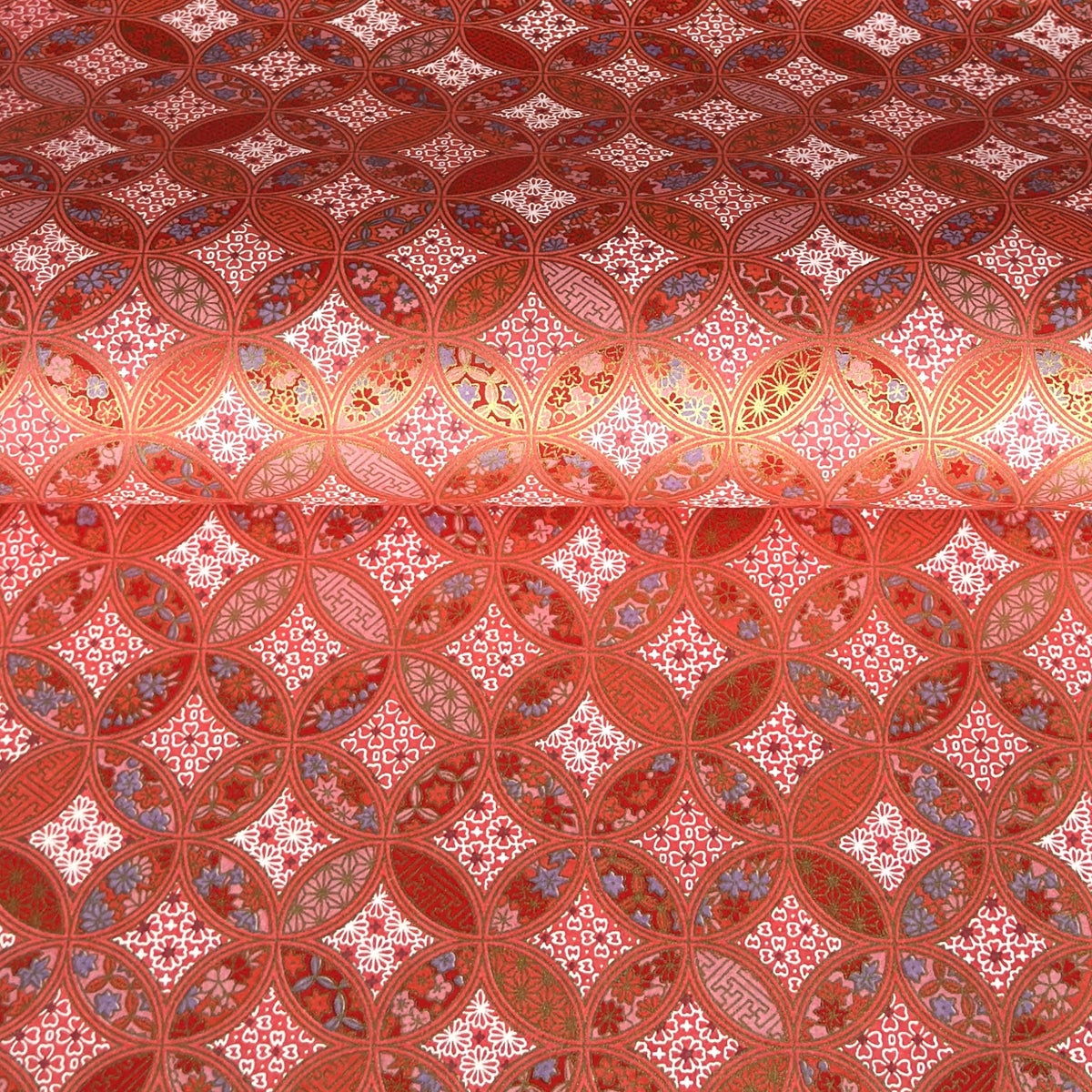 A close-up of a sheet of Japanese chiyogami paper featuring a repeating geometric medallion design in shades of terracotta red, coral pink, and metallic gold