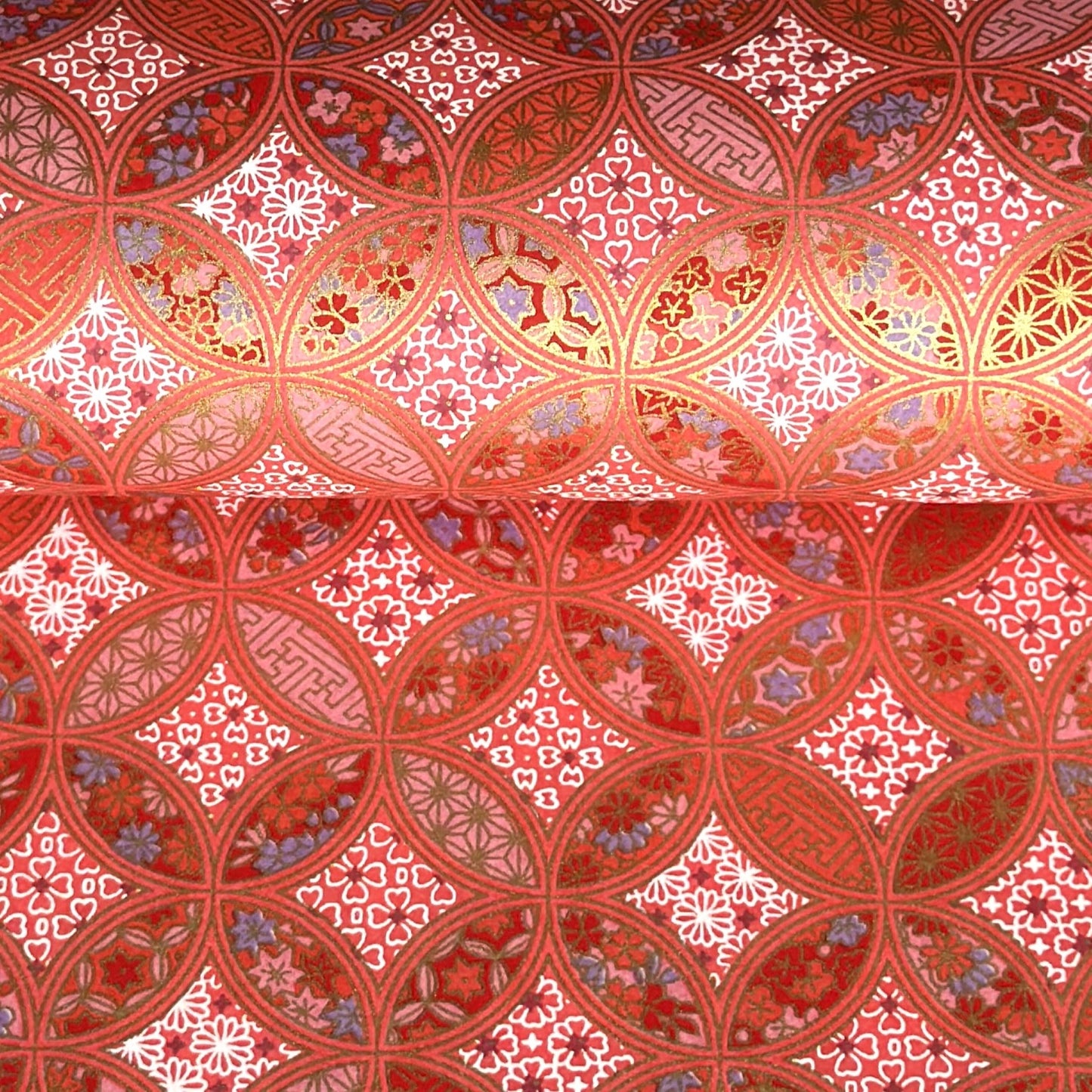 A close-up of a sheet of Japanese chiyogami paper featuring a repeating geometric medallion design in shades of terracotta red, coral pink, and metallic gold