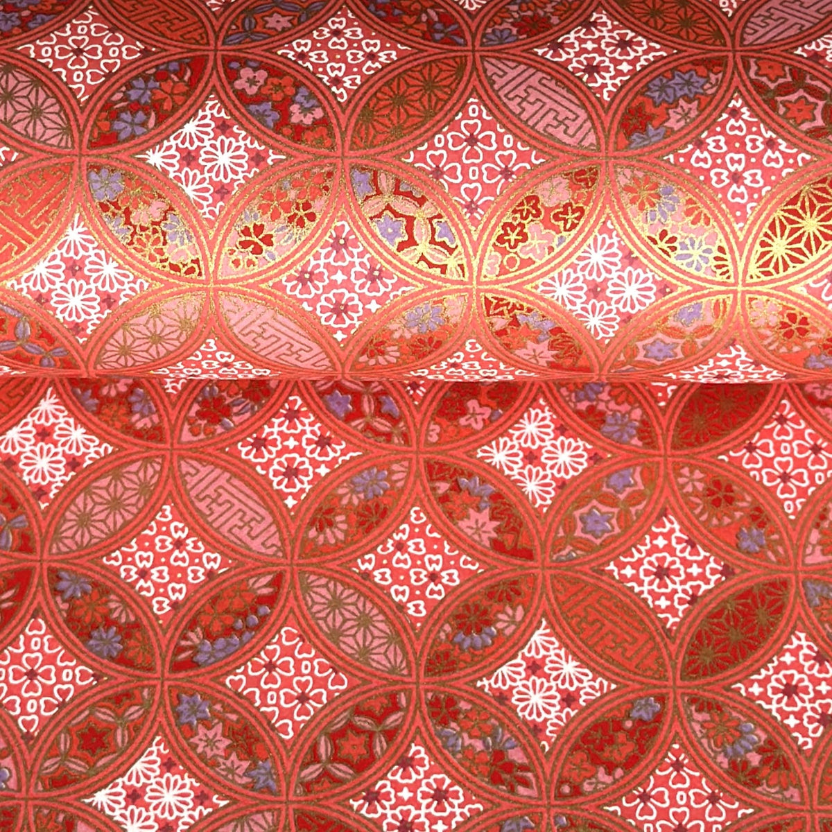 A close-up of a sheet of Japanese chiyogami paper featuring a repeating geometric medallion design in shades of terracotta red, coral pink, and metallic gold