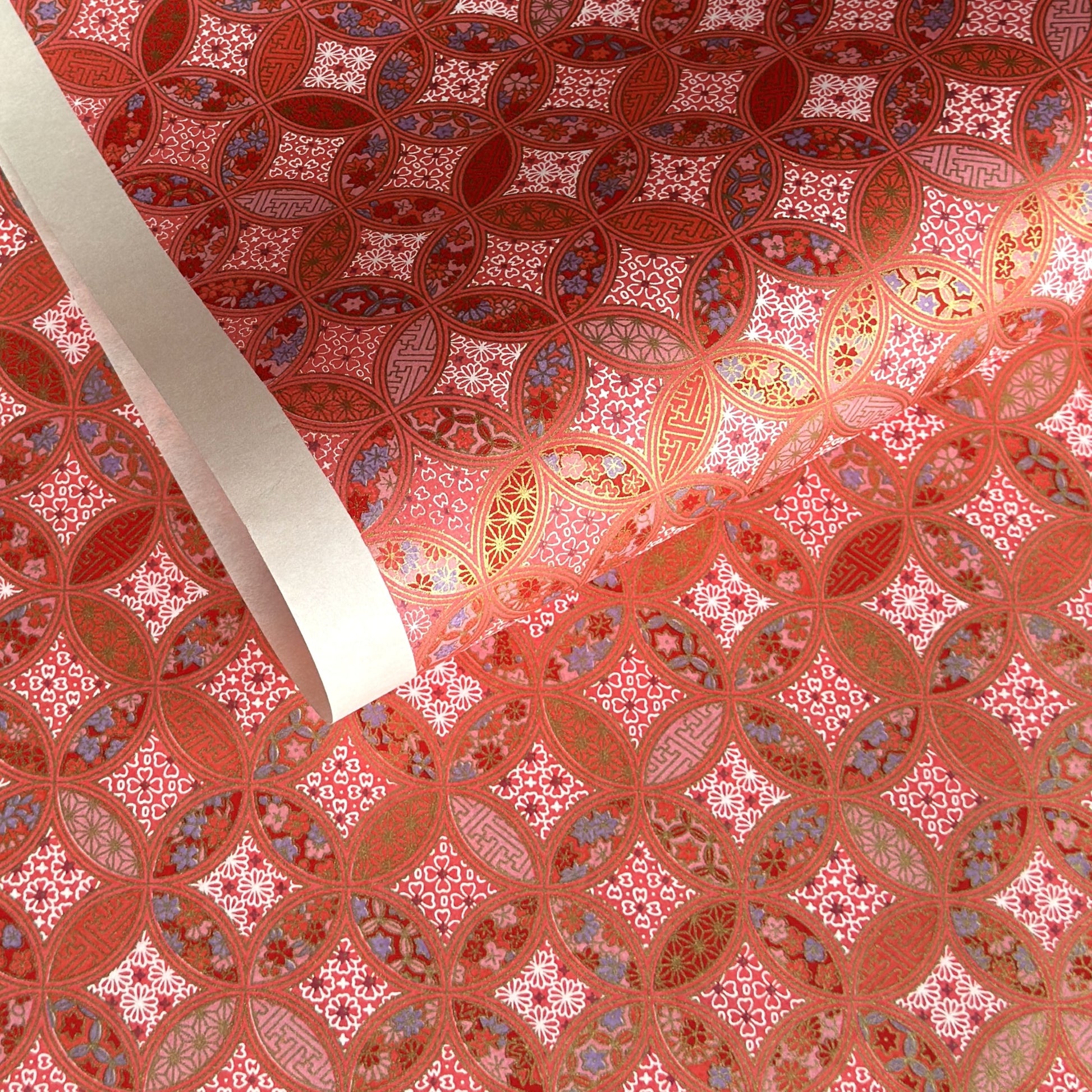 A close-up of a sheet of Japanese chiyogami paper featuring a repeating geometric medallion design in shades of terracotta red, coral pink, and metallic gold