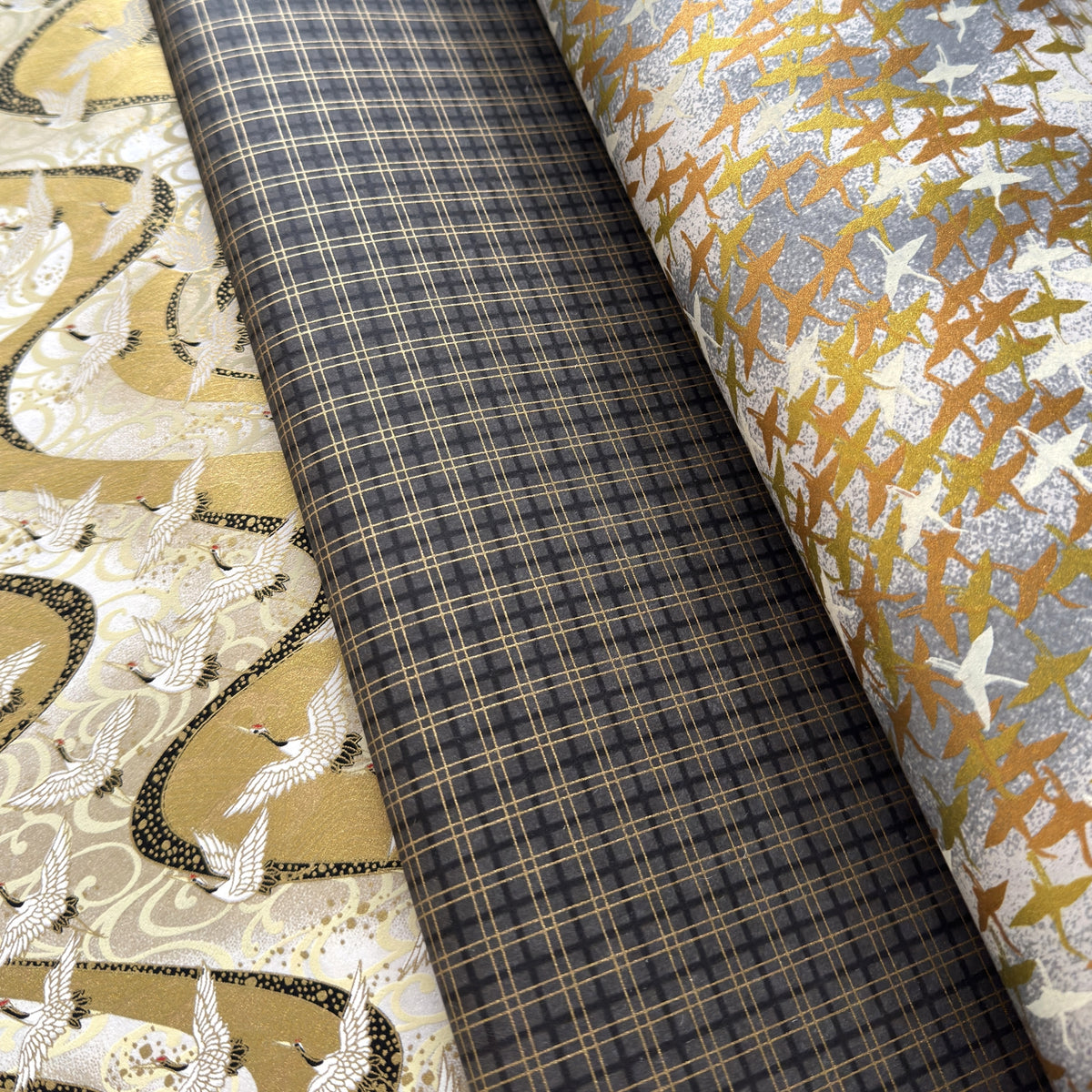 a sheet of Japanese chiyogami paper with a check plaid design in grey, black and gold.  alongside two other Japanese papers with patterns of gold birds