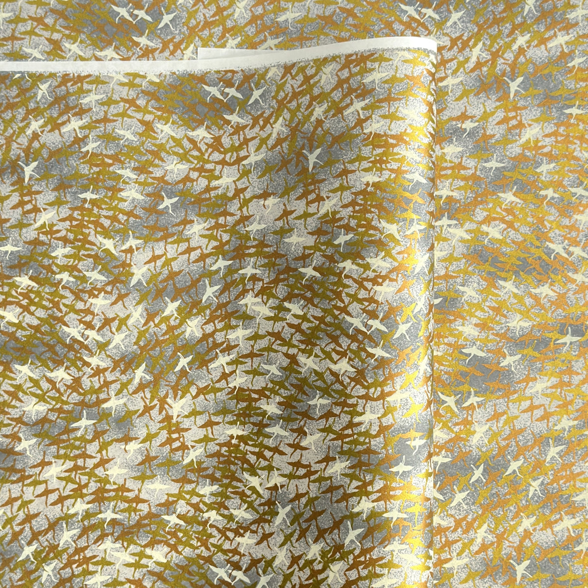 a sheet of Japanese yuzen chiyogami paper with a pattern of a flock of lots of birds flying. Birds are white, gold or copper in colour and flying on a ground of light grey and white sky