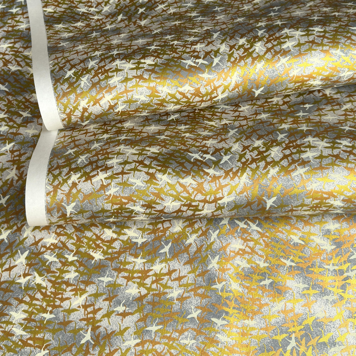 a sheet of Japanese yuzen chiyogami paper with a pattern of a flock of lots of birds flying. Birds are white, gold or copper in colour and flying on a ground of light grey and white sky