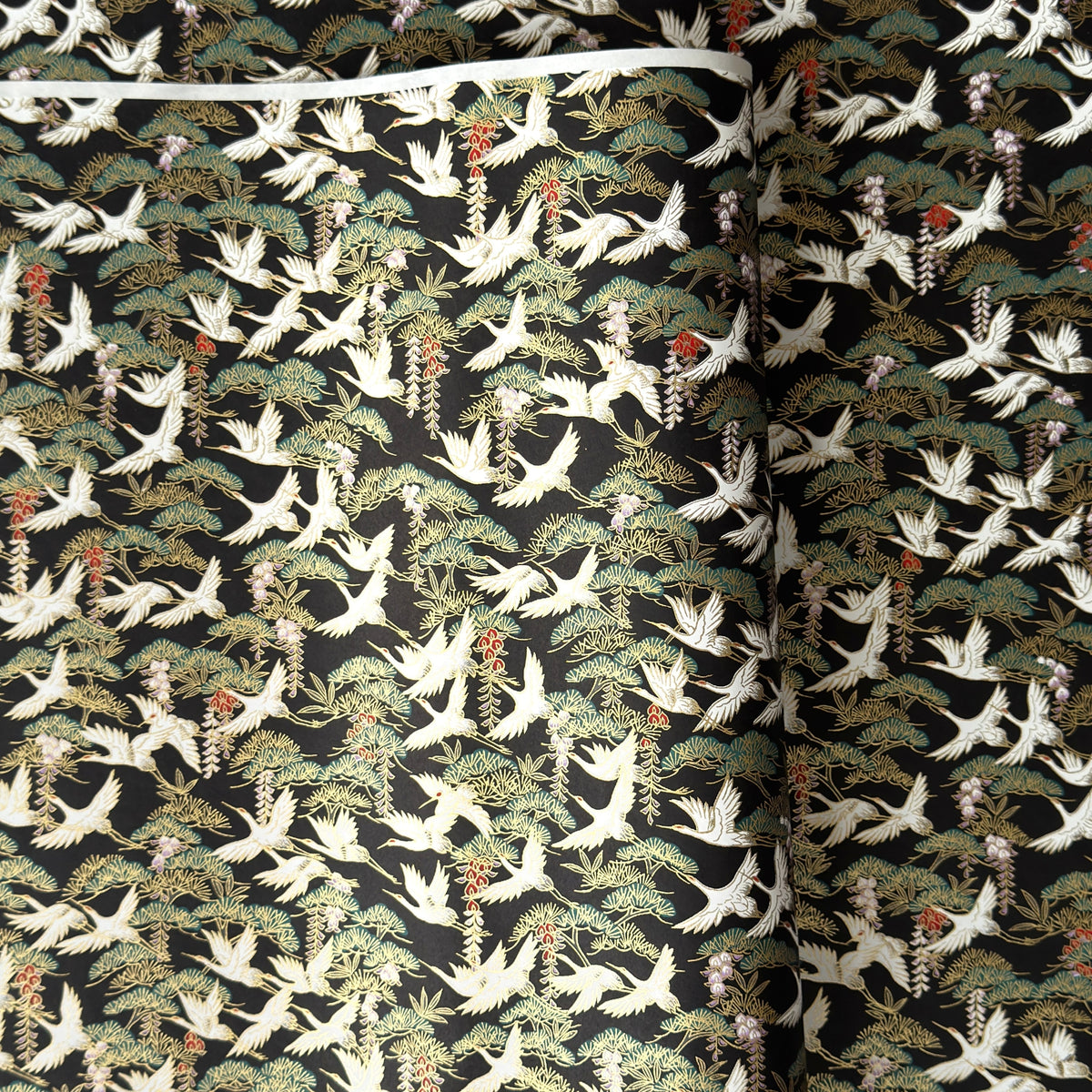 A close-up of a sheet of Japanese chiyogami paper featuring white cranes flying over green pine trees and branches on a black background.