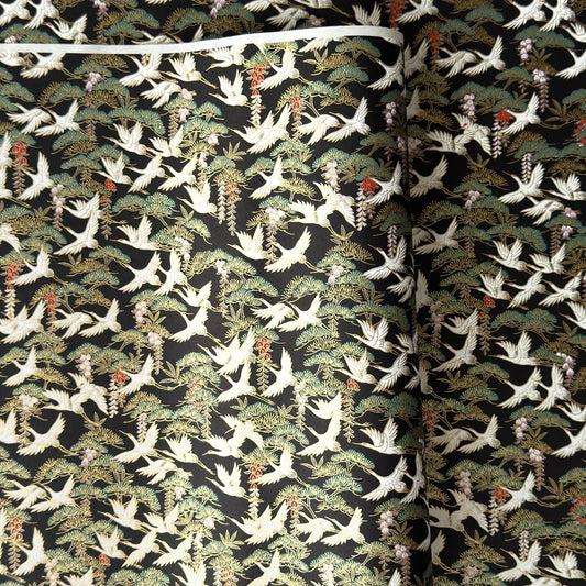A close-up of a sheet of Japanese chiyogami paper featuring white cranes flying over green pine trees and branches on a black background.