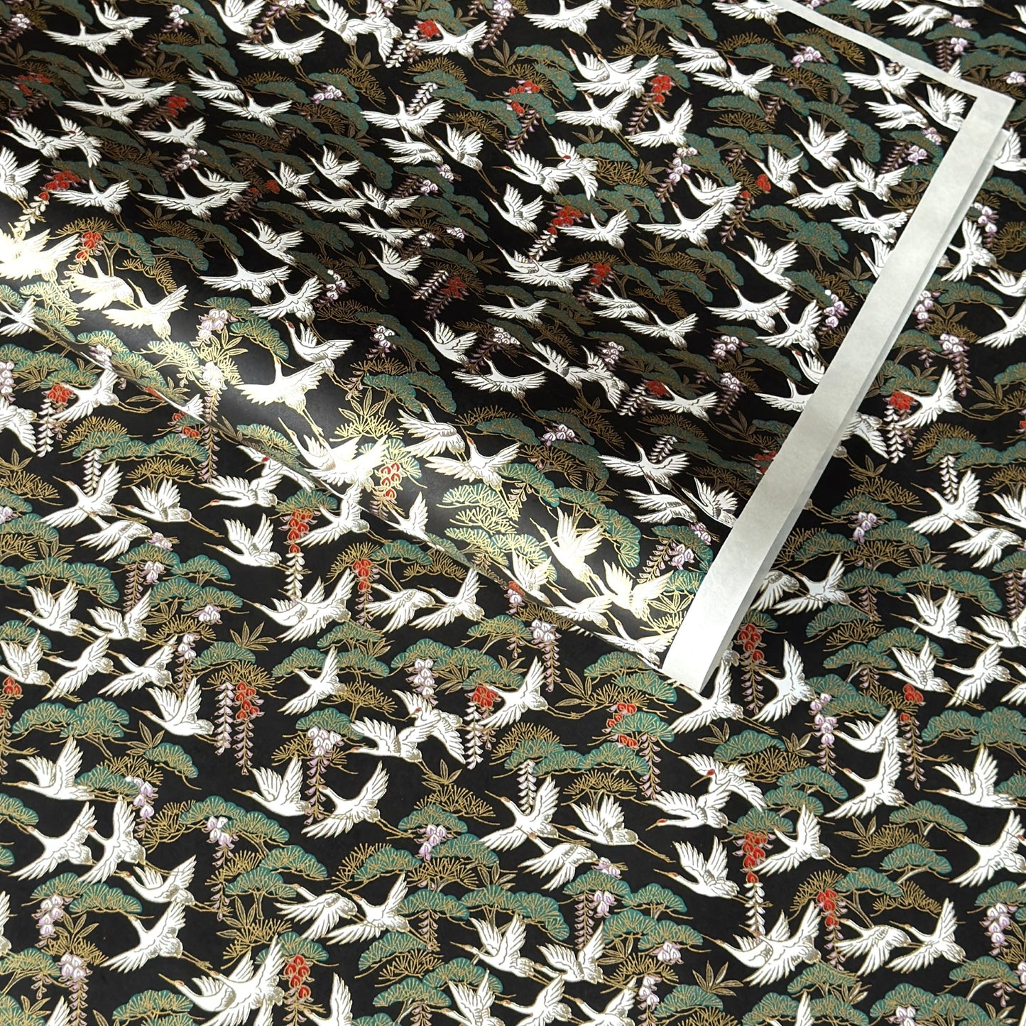 A close-up of a sheet of Japanese chiyogami paper featuring white cranes flying over green pine trees and branches on a black background.