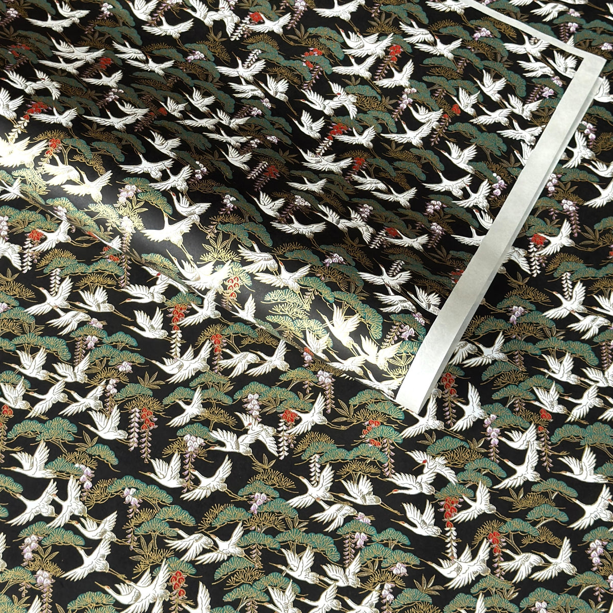 A close-up of a sheet of Japanese chiyogami paper featuring white cranes flying over green pine trees and branches on a black background.