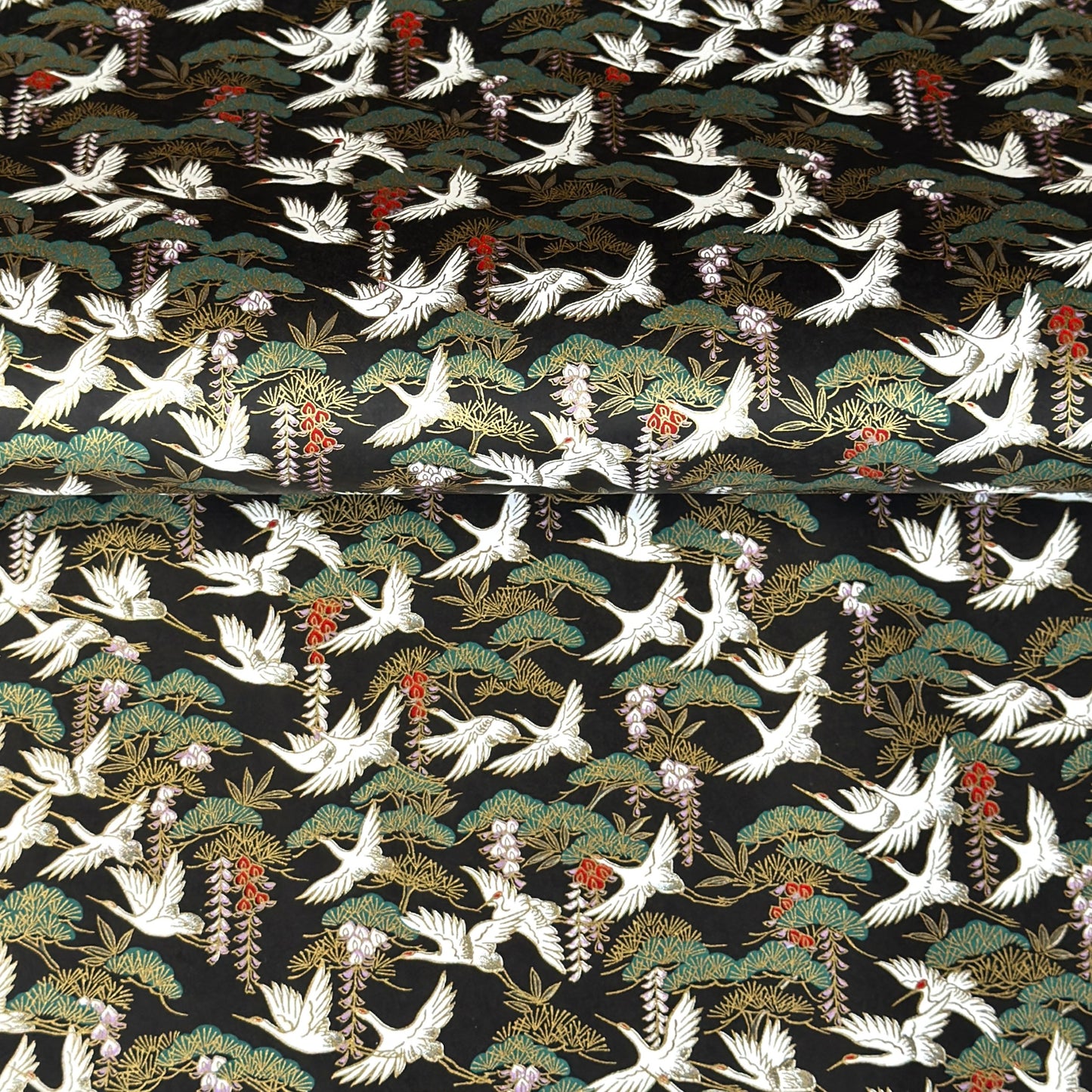 A close-up of a sheet of Japanese chiyogami paper featuring white cranes flying over green pine trees and branches on a black background.